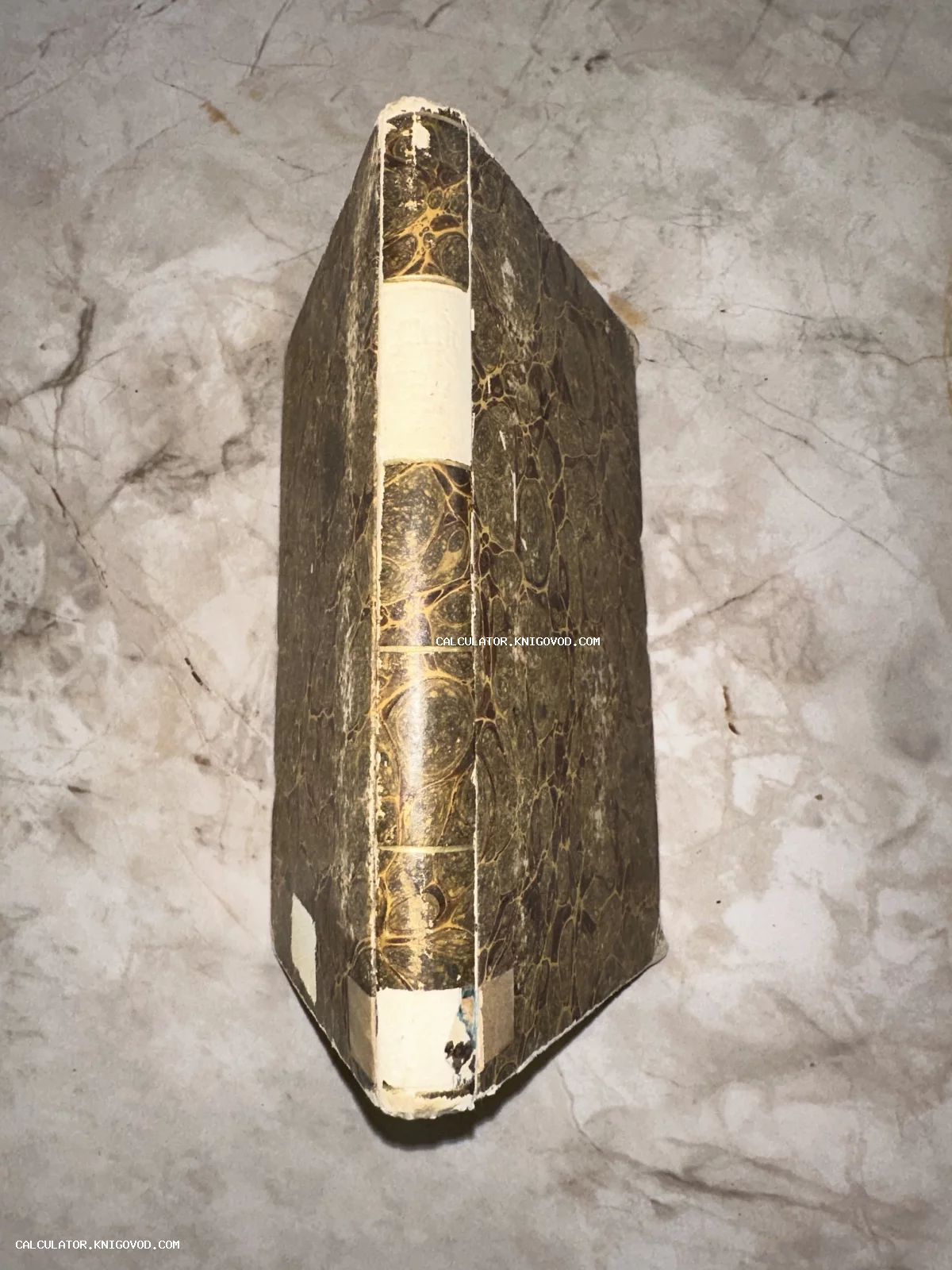 Вид сверху на корешок и обложку старинной книги с характерным мраморным узором бумаги на фоне светлой каменной поверхности.