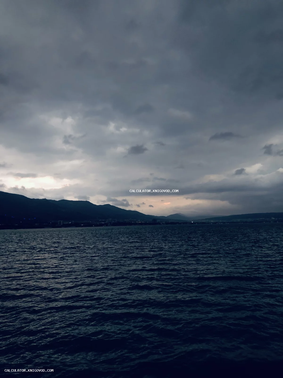 Вид на темную водную гладь и силуэты гор под тяжелыми грозовыми облаками в сумерках.