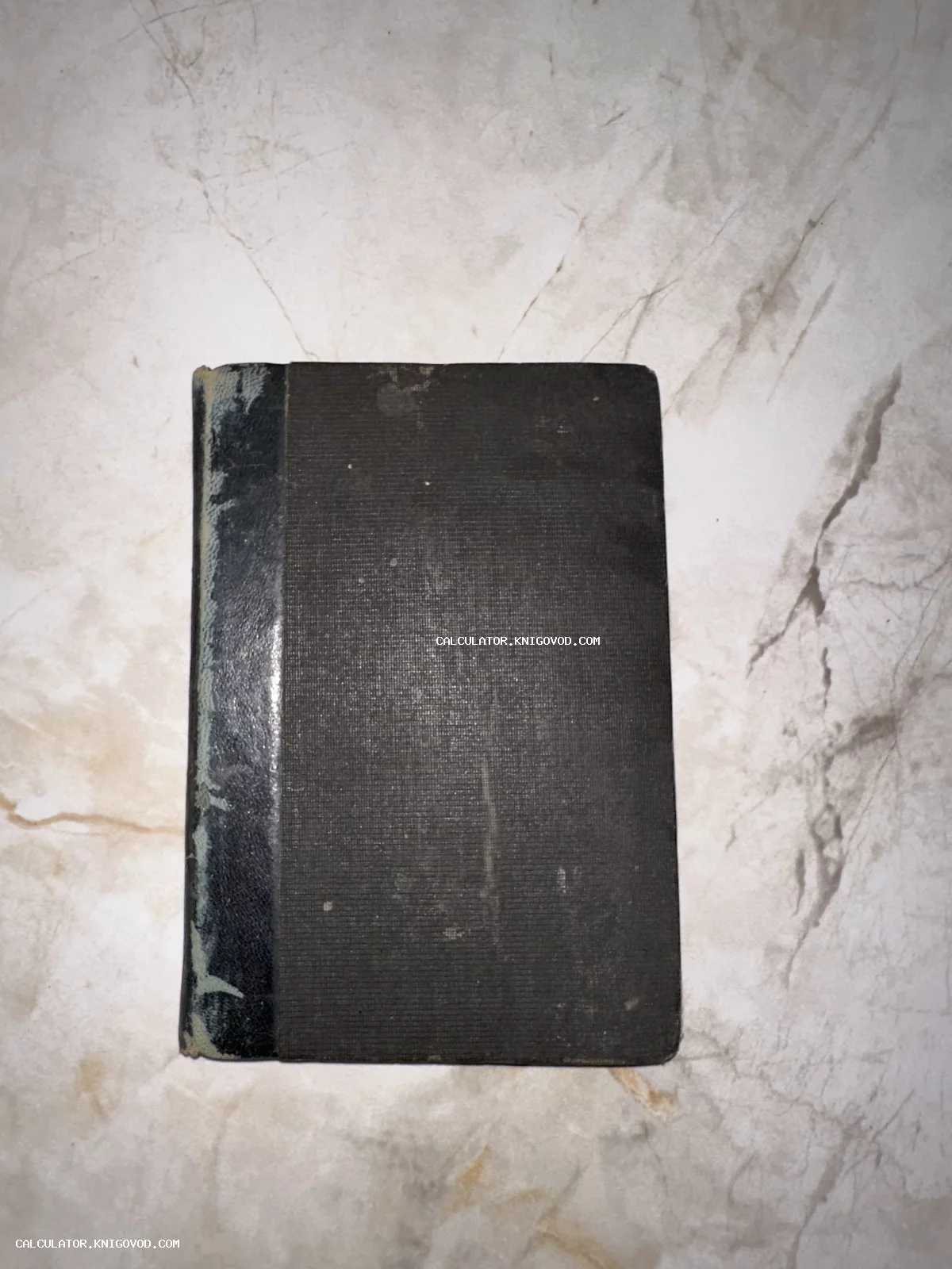 Старинная книга в черном тканевом переплете с потертым корешком на светлой мраморной поверхности.