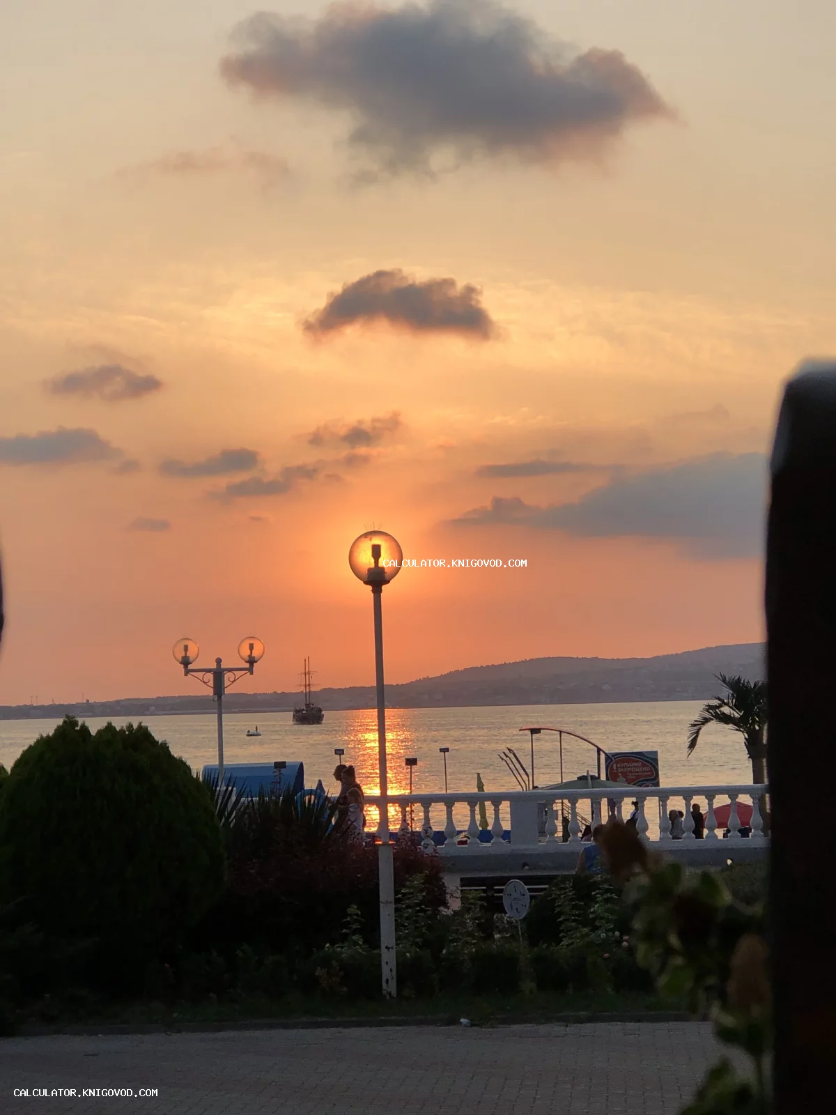 Фотография заката на морской набережной: солнце совпадает с плафоном уличного фонаря, на фоне моря и гор виден парусник.