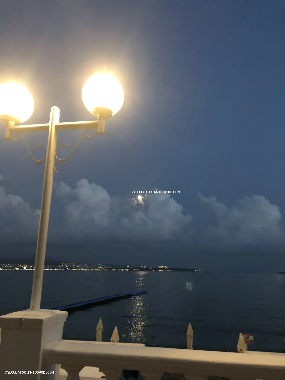 Классический белый уличный фонарь с двумя круглыми плафонами на фоне ночного моря и облачного неба с луной.