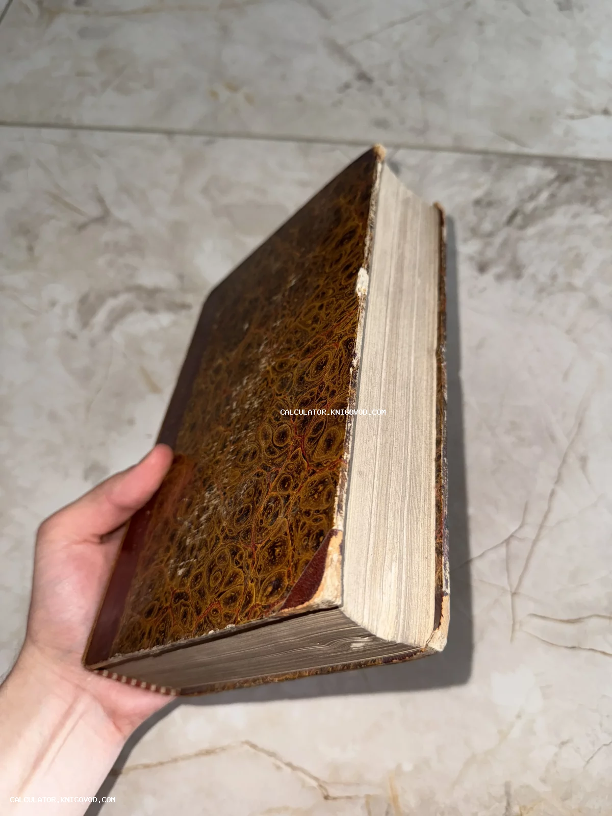 Старинная толстая книга с обложкой под мрамор в руке человека на фоне светлого пола.