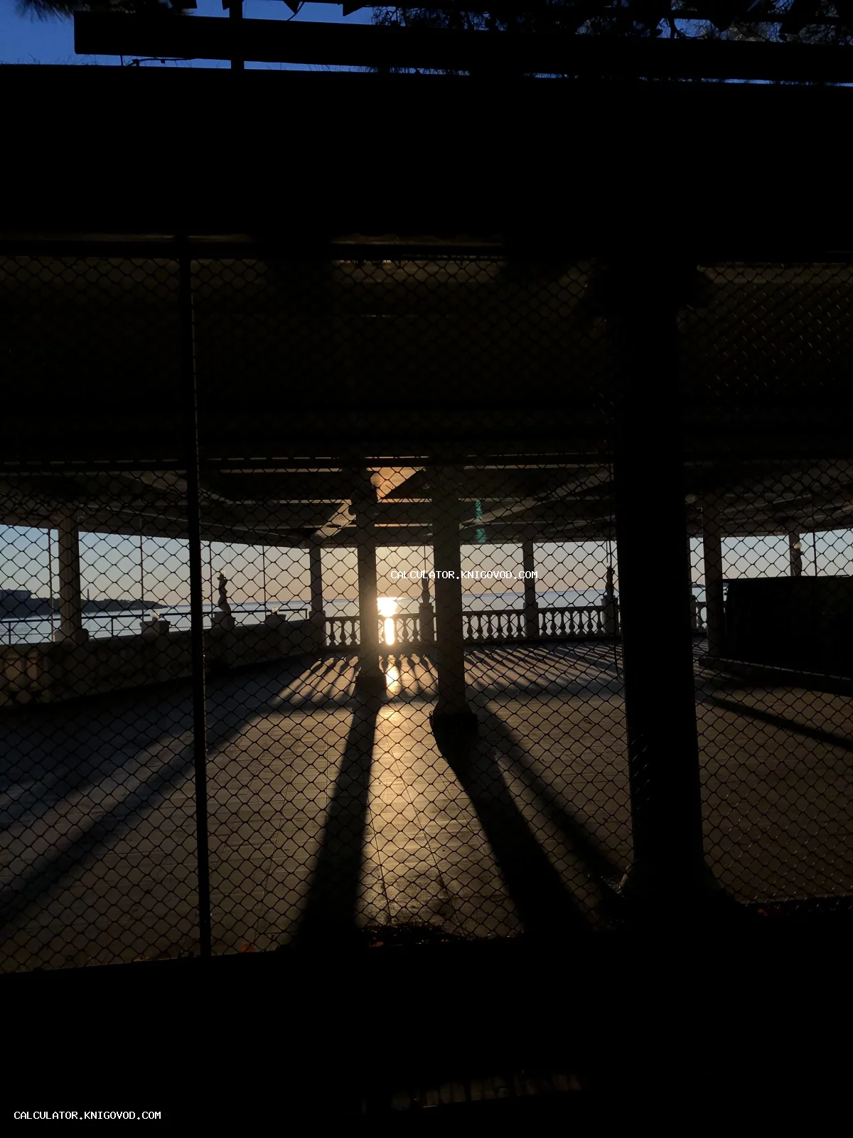 Вид на закатное солнце над морем через металлическую сетку и колонны классического павильона.