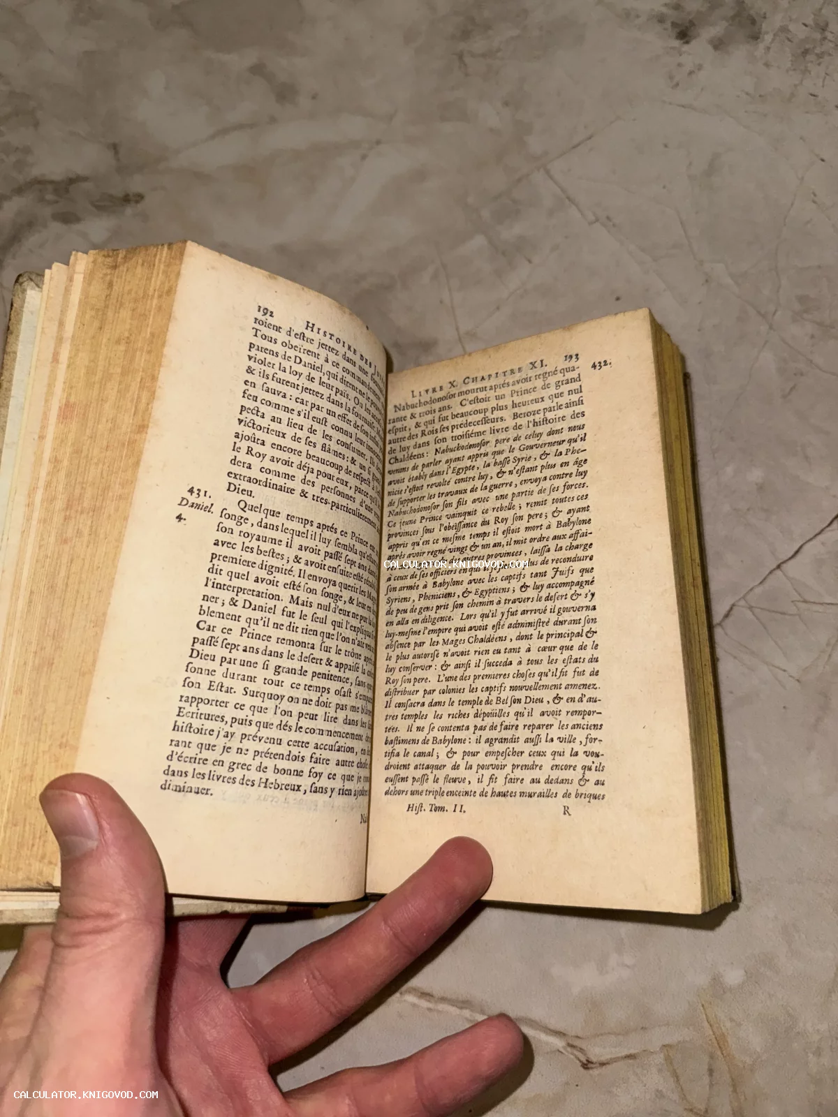 Раскрытая старинная книга с пожелтевшими страницами и французским текстом, которую держит рука человека на фоне мраморного пола.