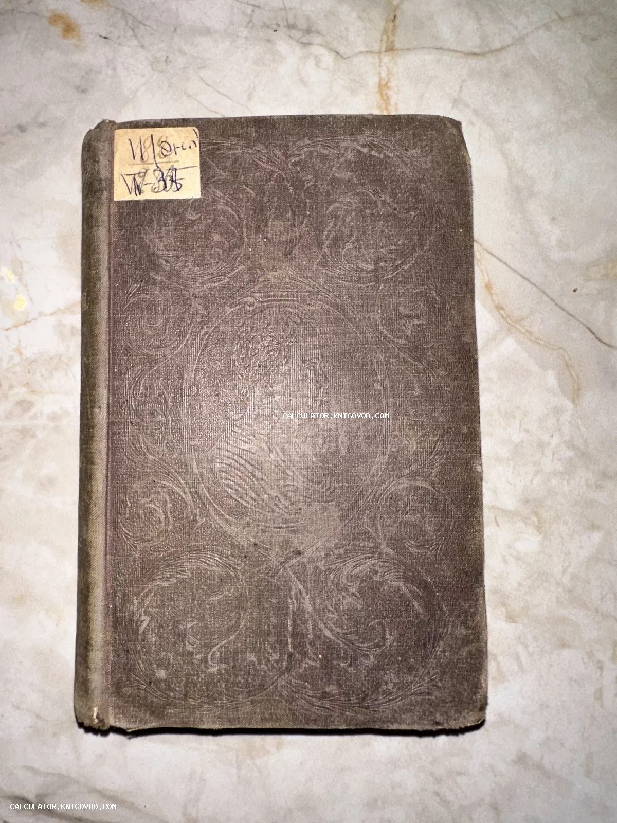 Вид сверху на обложку старинной антикварной книги коричневого цвета с тисненым растительным орнаментом и бумажной наклейкой с рукописным номером.