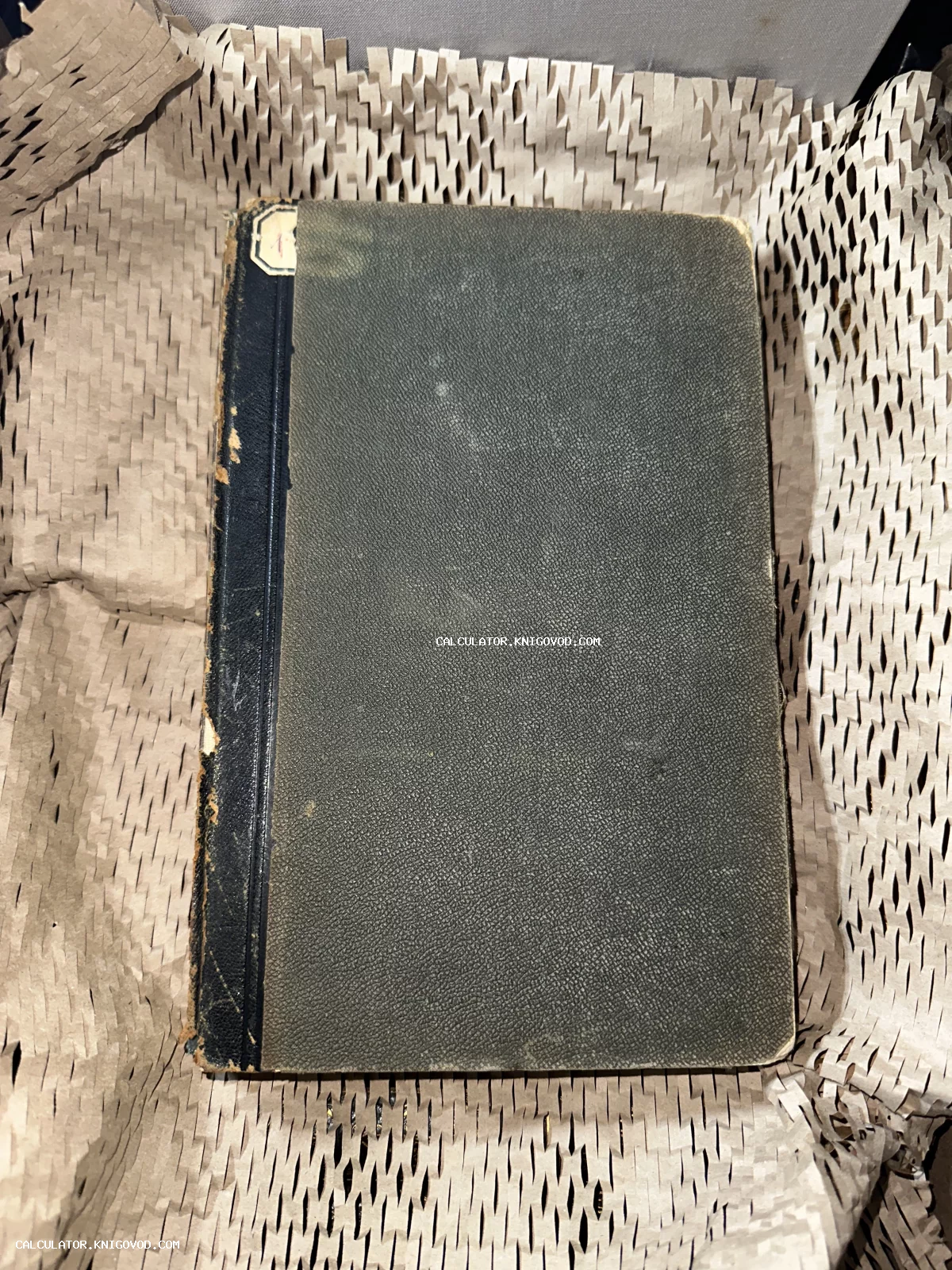 Старинная книга с темной фактурной обложкой и потертым синим корешком, лежащая на упаковочной бумаге.