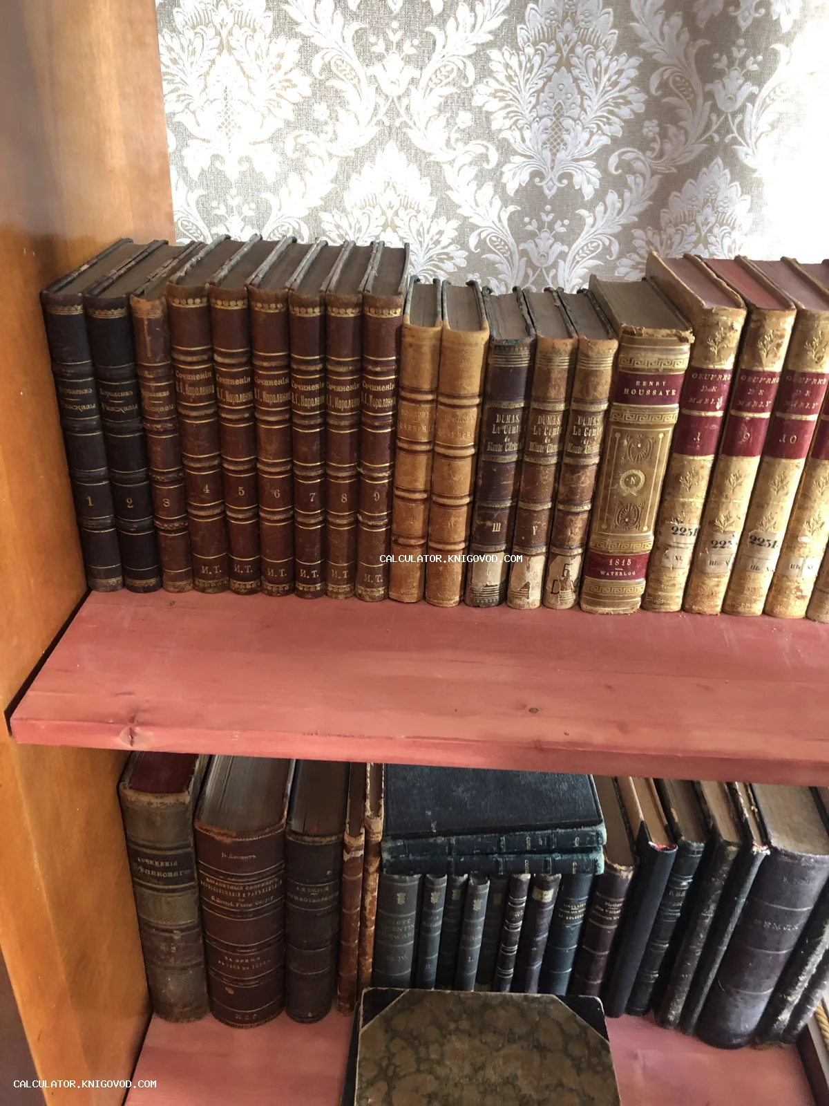 Старинные книги с золотым тиснением на корешках, стоящие на деревянной полке на фоне узорчатых обоев.