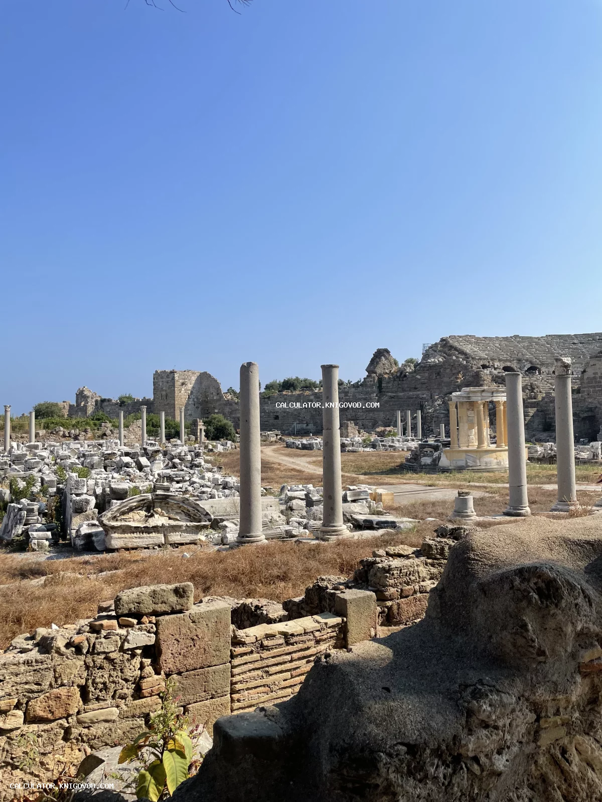 Античные каменные колонны, фрагменты стен и остатки древних построек под ясным синим небом.