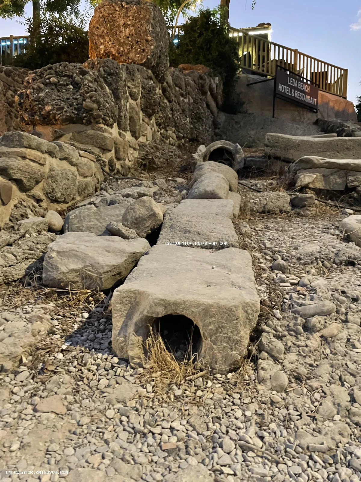 Древние каменные блоки с круглыми отверстиями внутри, уложенные в ряд и представляющие собой фрагменты античной системы водоснабжения на фоне руин.