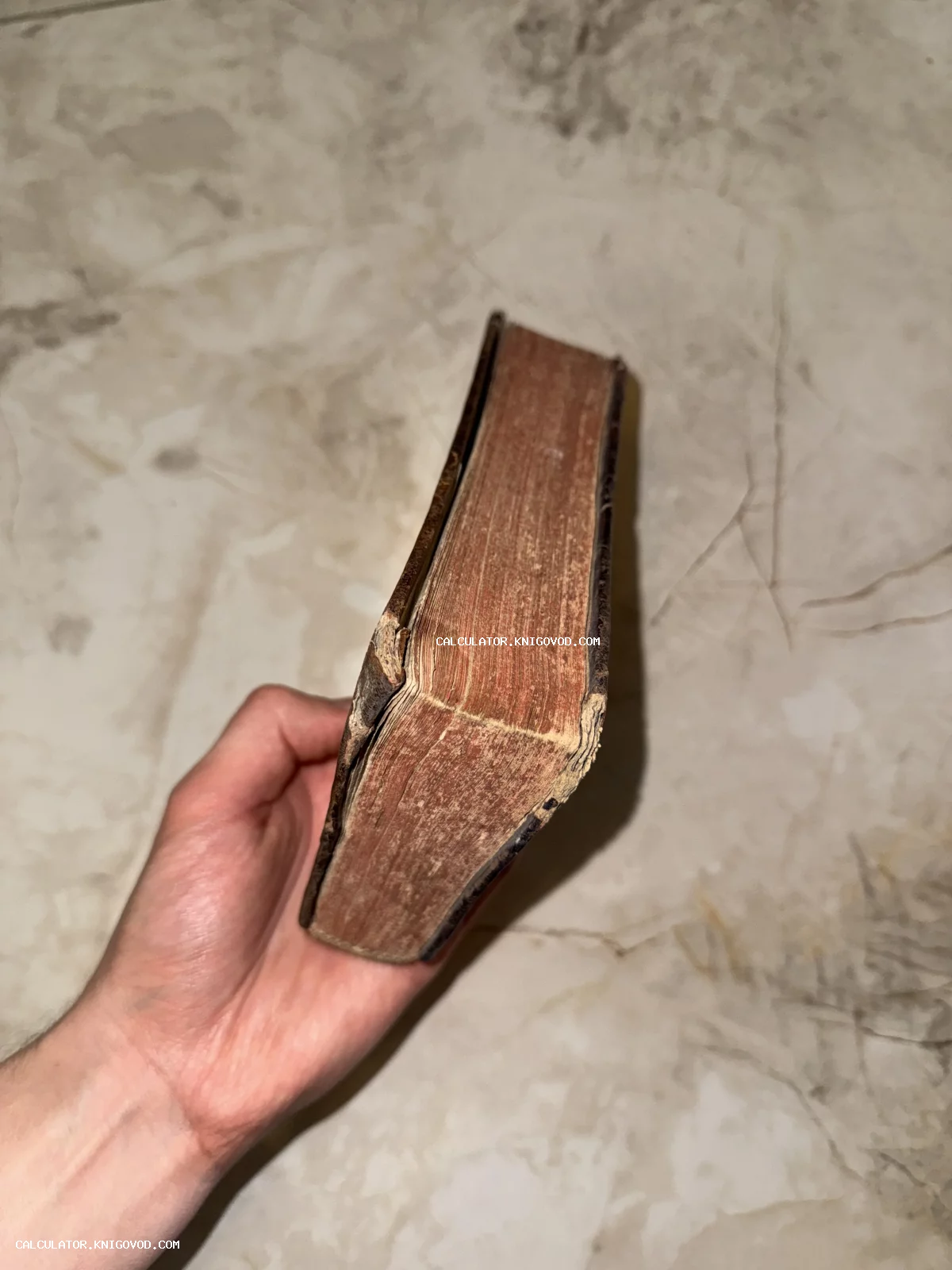 Вид сбоку на массивный блок страниц старинной книги с потертым кожаным переплетом в руке человека.