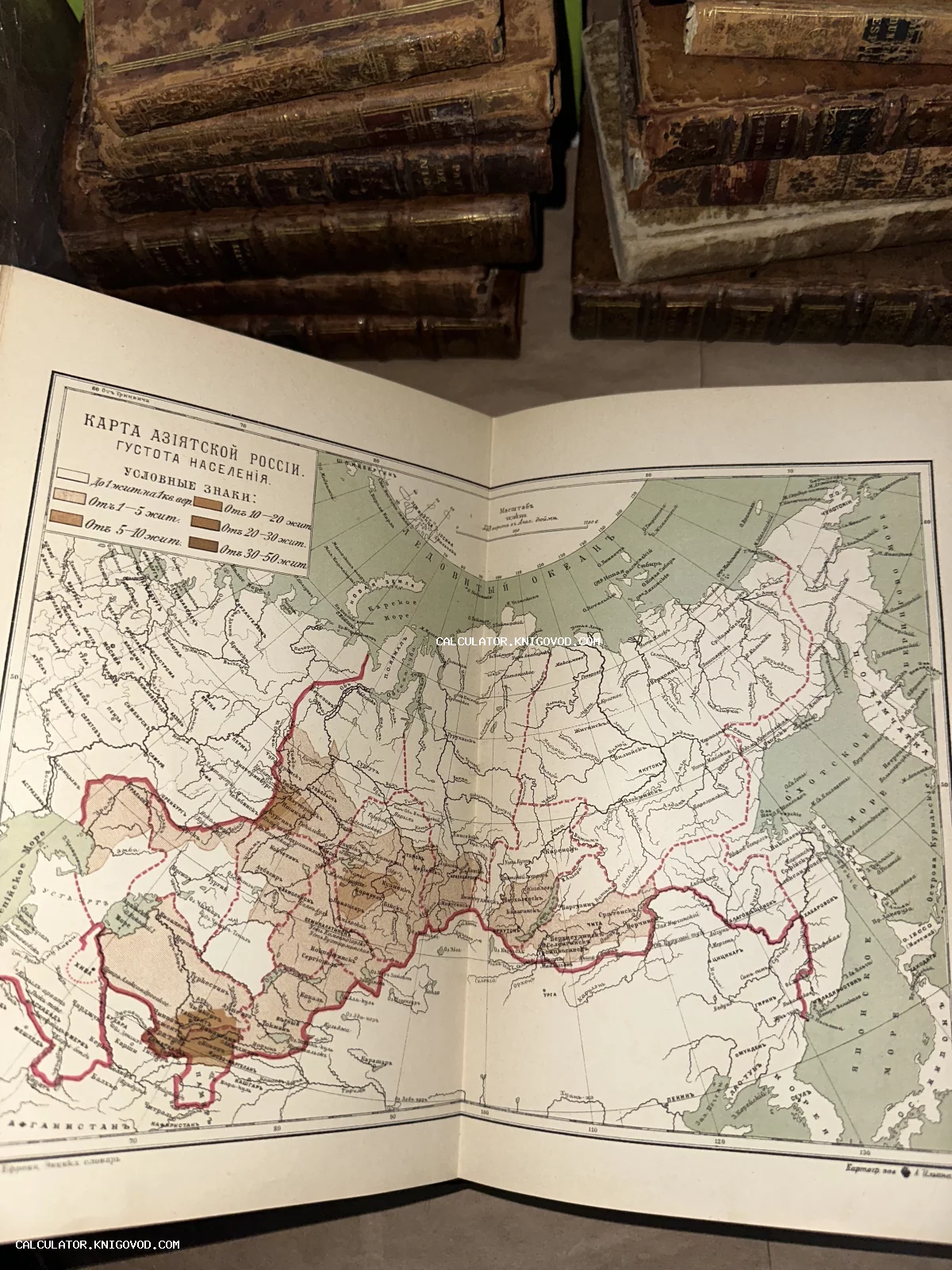 Разворот антикварной книги с картой плотности населения Азиатской России на фоне стопок старых книг в кожаных переплетах.