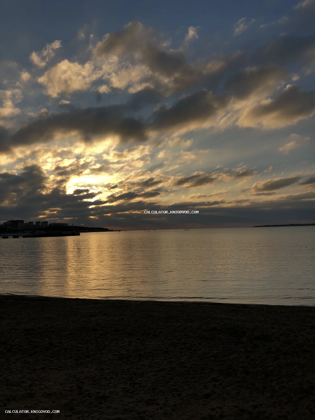 Морской пейзаж на закате: темный песчаный пляж на переднем плане, спокойная гладь воды с солнечной дорожкой и облачное небо с пробивающимися лучами.
