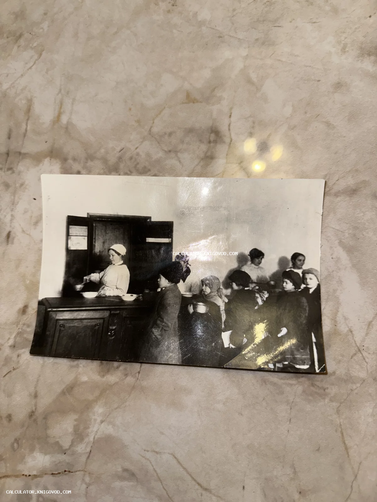 Черно-белый исторический снимок: женщина в белом фартуке и головном уборе разливает еду детям из большой кастрюли в столовой.