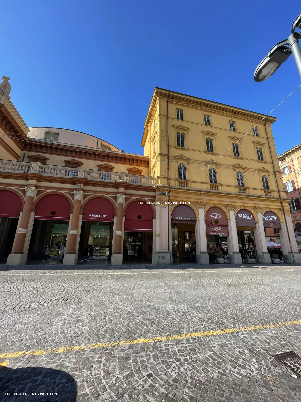 Вид на историческое здание с высокими арочными галереями и брусчаткой на площади под ярким синим небом.