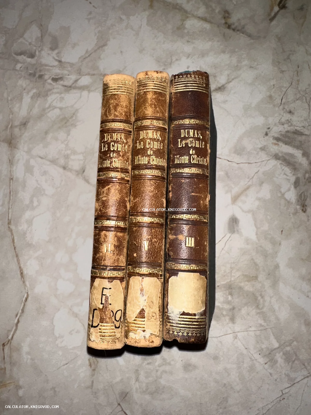 Корешки трех томов антикварной книги Александра Дюма «Le Comte de Monte-Christo» в кожаном переплете с золотым тиснением на мраморном фоне.