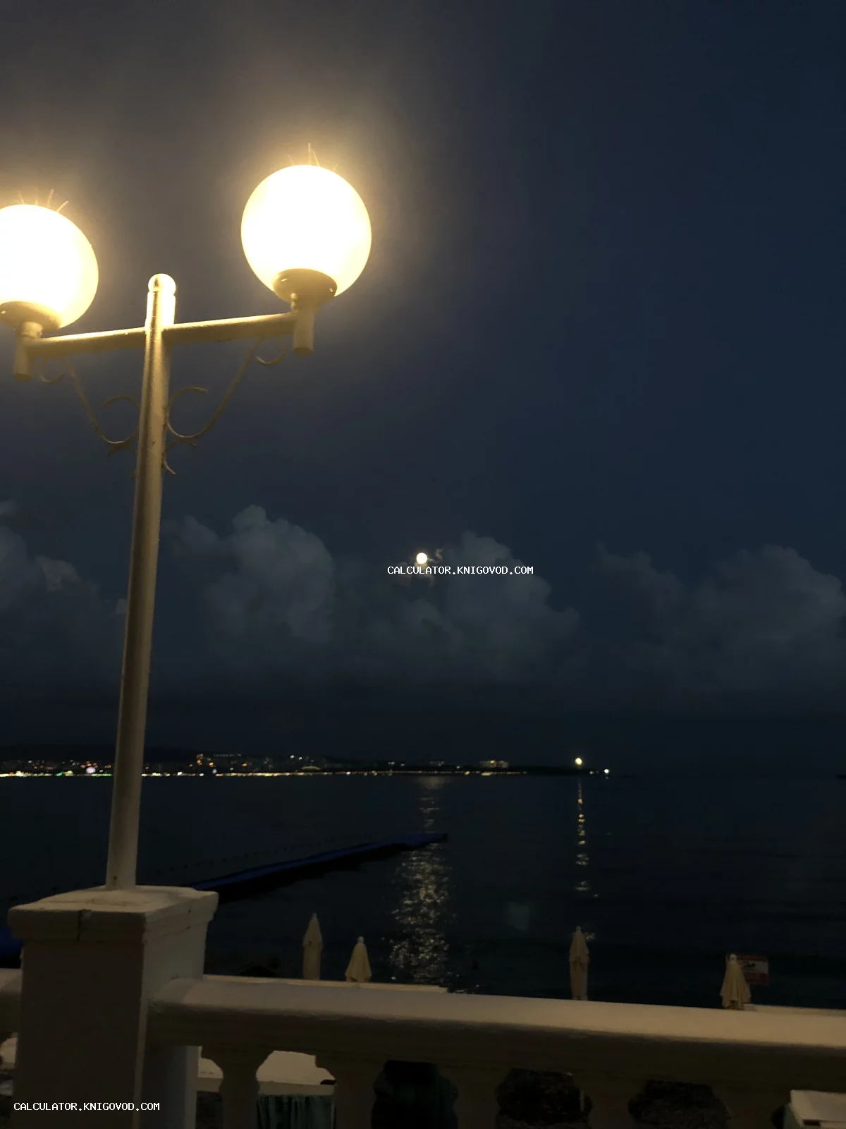 Уличный фонарь с двумя светящимися круглыми плафонами на фоне ночного моря, луны и облаков.