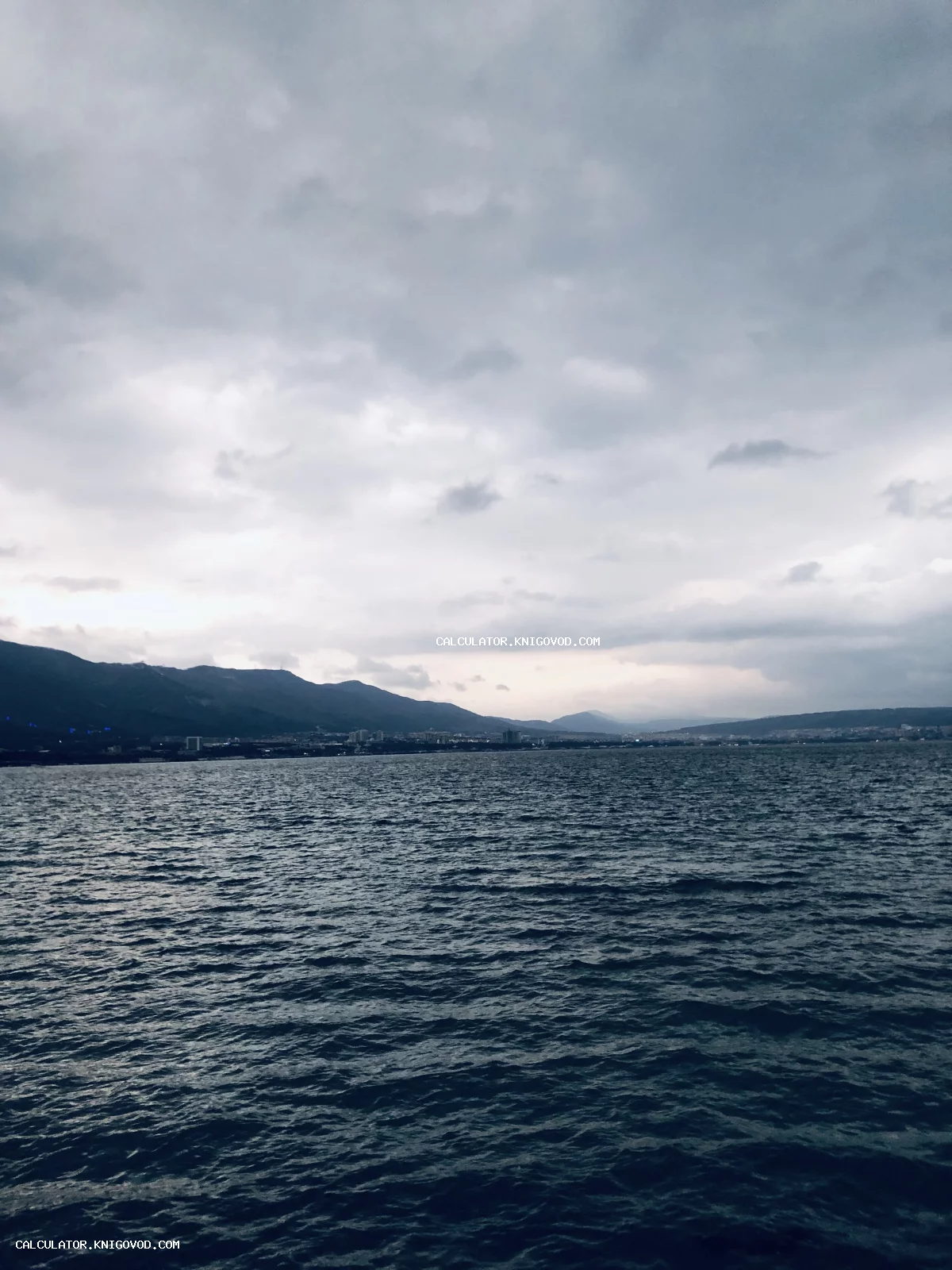 Темная морская гладь с мелкой рябью, на заднем плане очертания горного хребта и пасмурное небо в облаках.