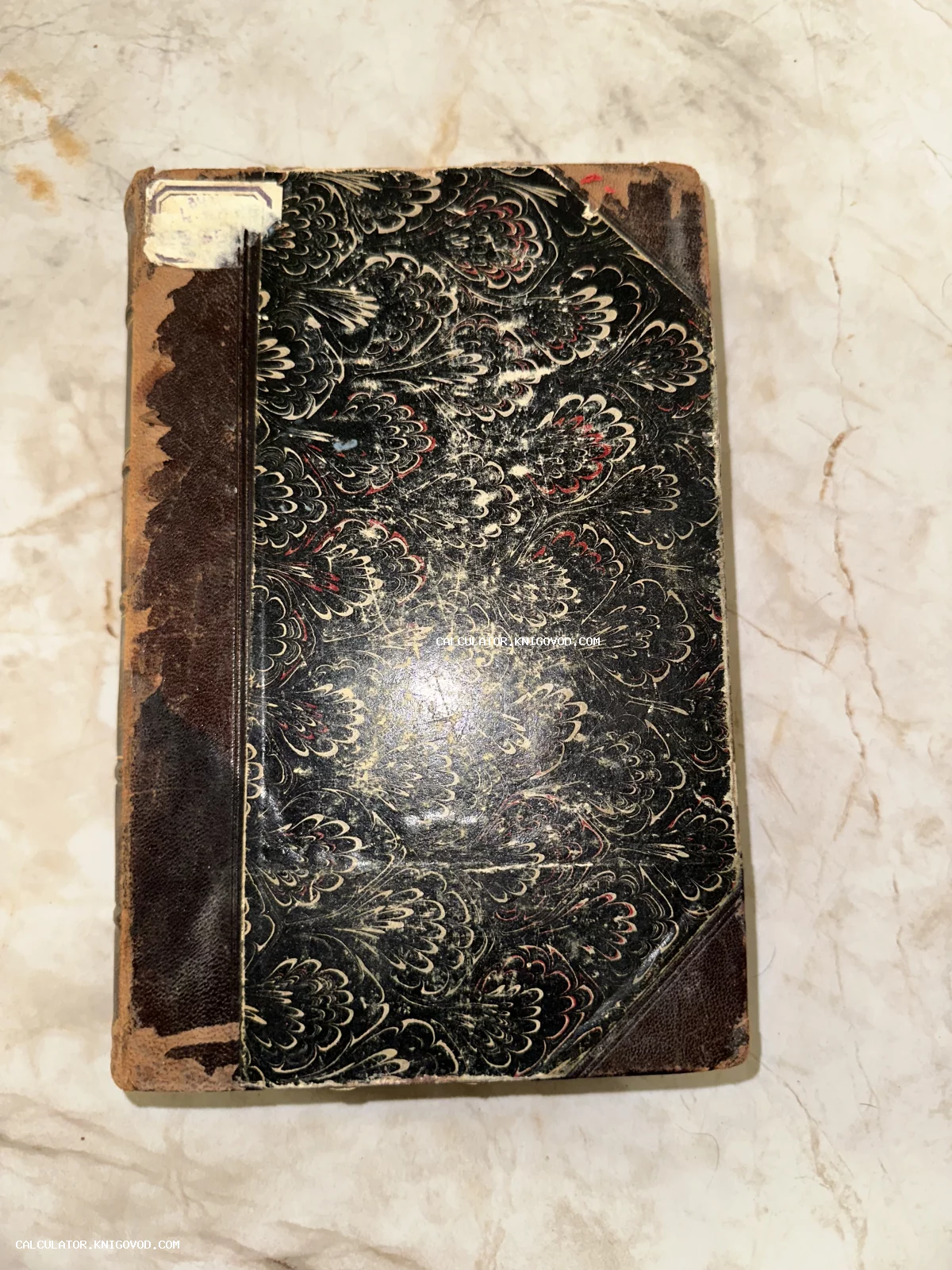 Старинная книга с черно-белым мраморным узором на обложке и кожаным корешком на светлой поверхности.