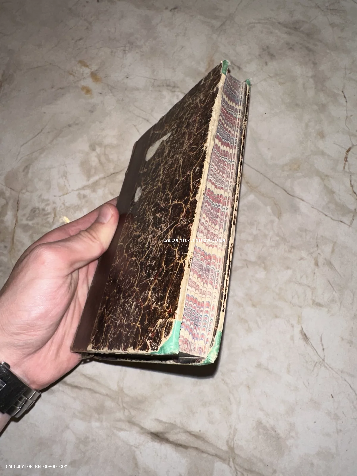 Человек держит в руке старинную антикварную книгу в потертом переплете с цветным декоративным мраморным обрезом.