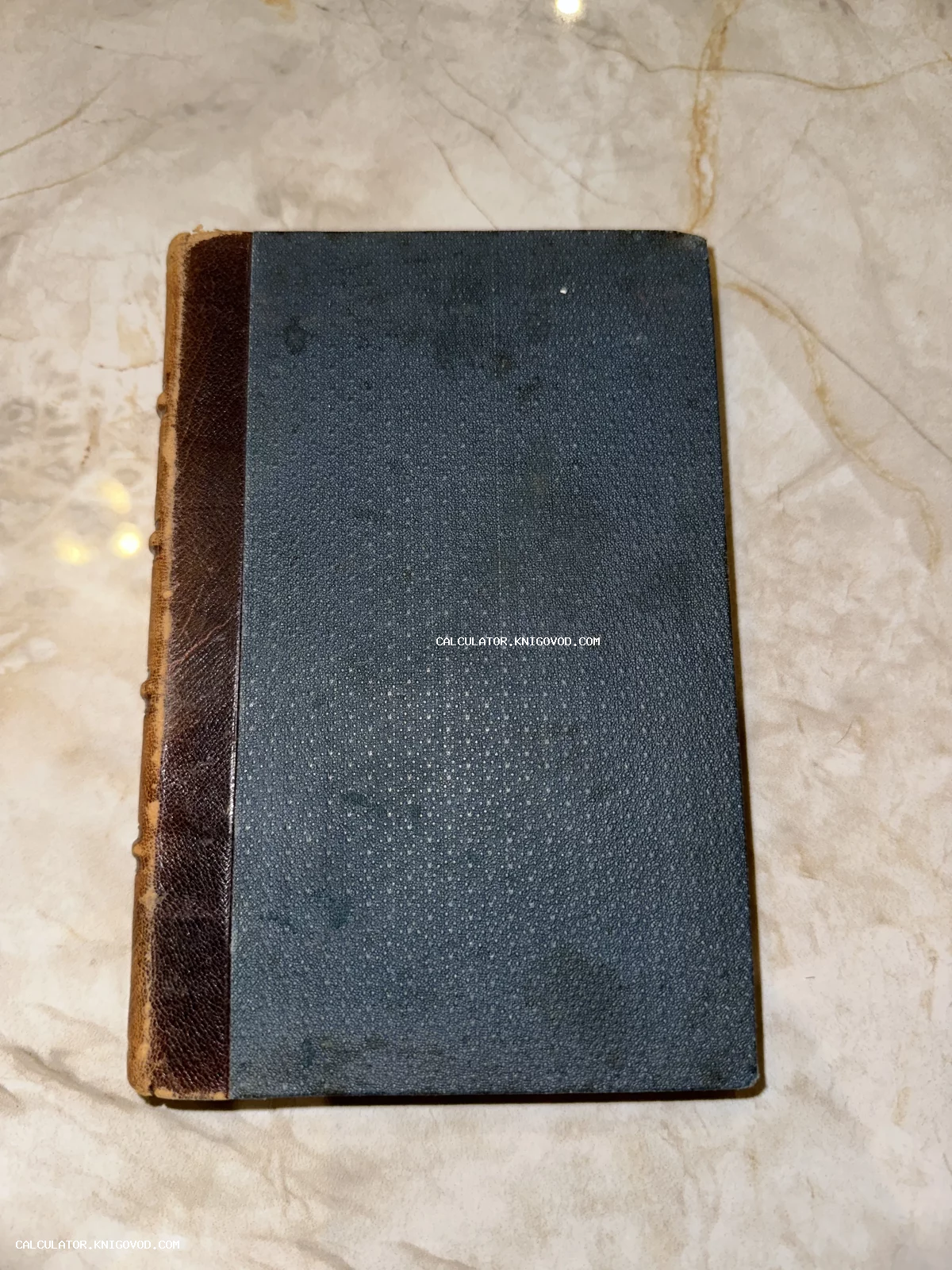 Старинная книга с темно-синей фактурной обложкой и коричневым кожаным корешком на светлой мраморной поверхности.