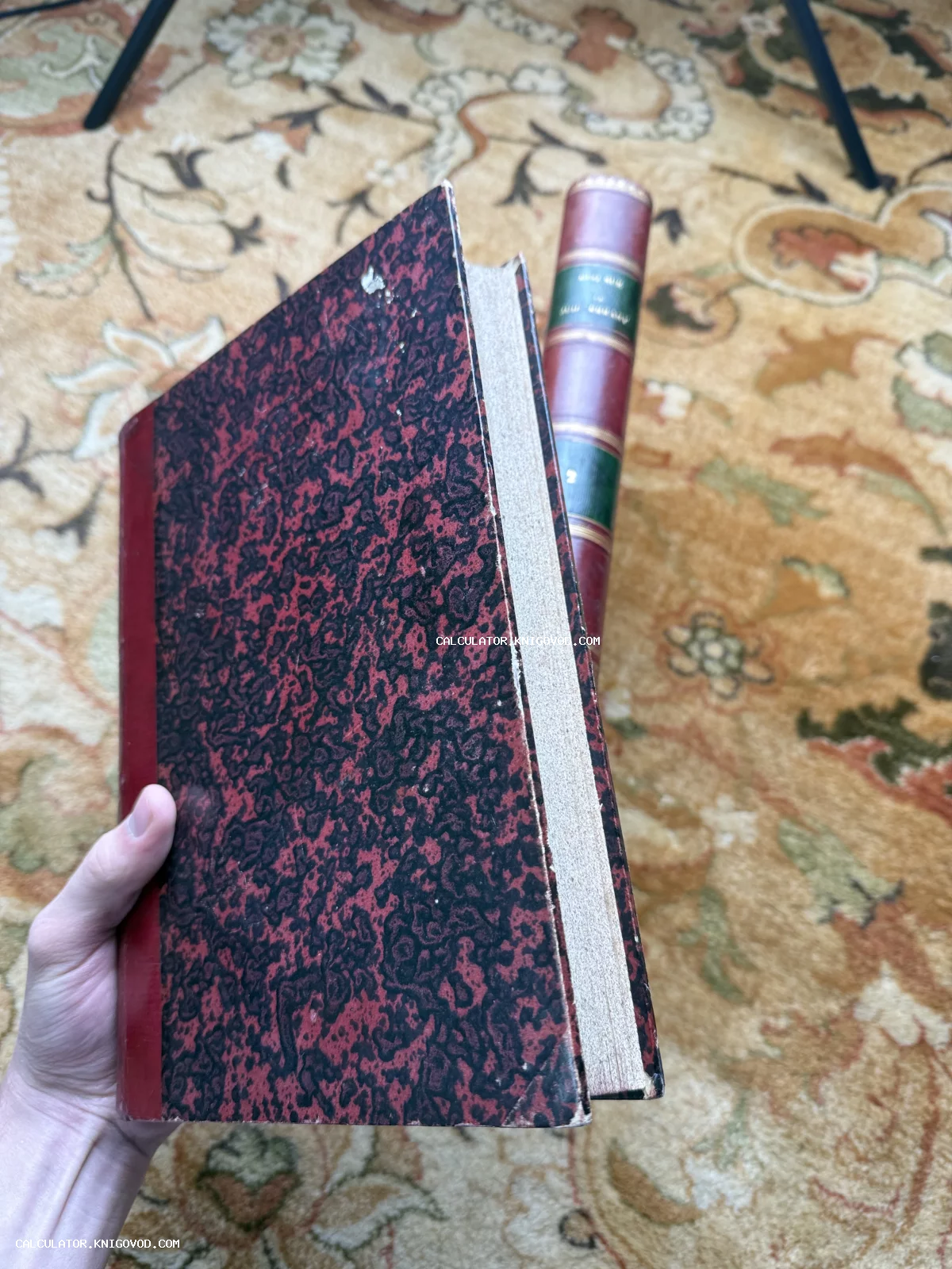 Старинная книга с красно-черной мраморной обложкой и красным кожаным корешком в руках на фоне ковра.