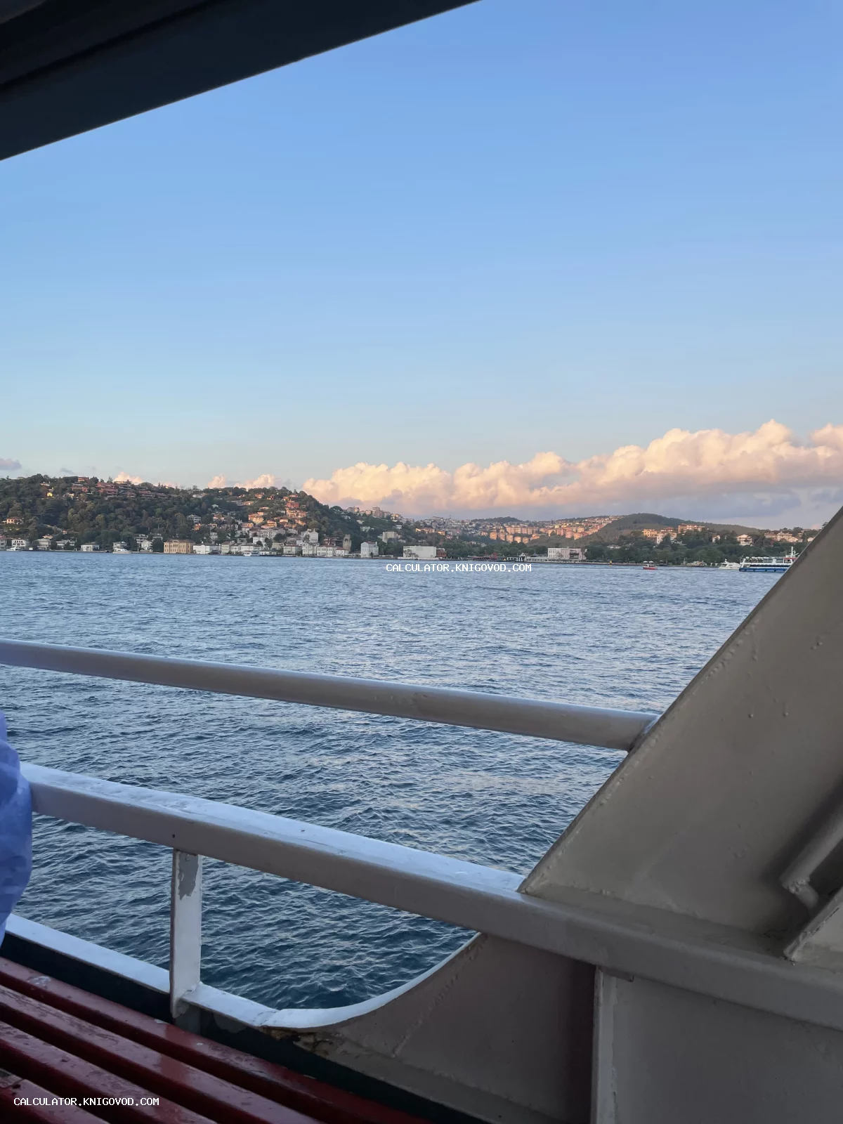 Вид на морское побережье с домами на холмах, снятый через перила палубы корабля в предзакатный час