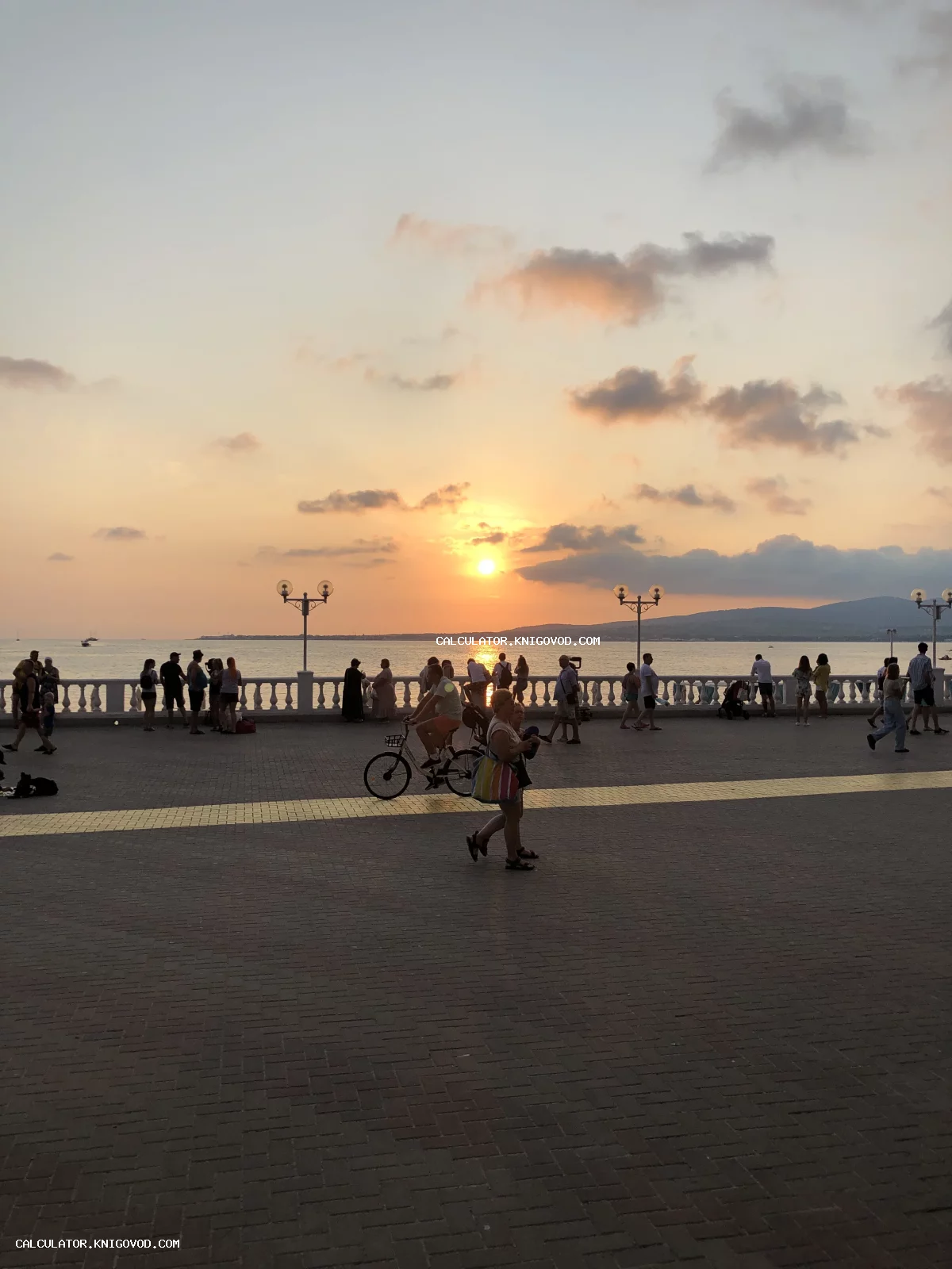 Силуэты людей на набережной с классической балюстрадой на фоне заходящего над морем солнца.