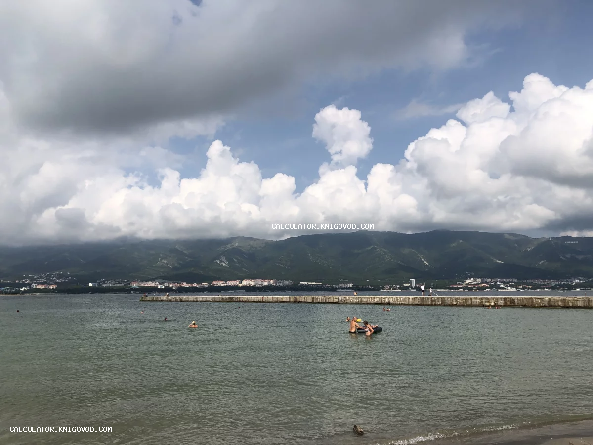 Вид на море, бетонный волнорез и горную гряду под густыми белыми облаками, люди купаются в воде.