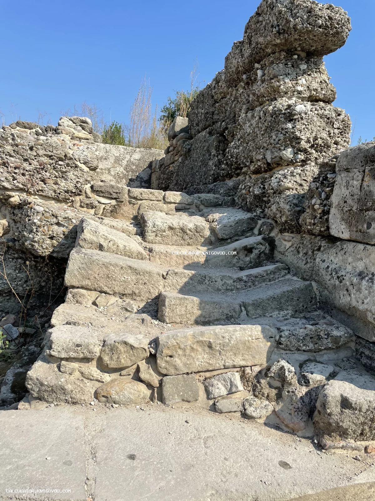 Вид снизу на широкие ступени из грубого камня, ведущие к полуразрушенным стенам античного сооружения.