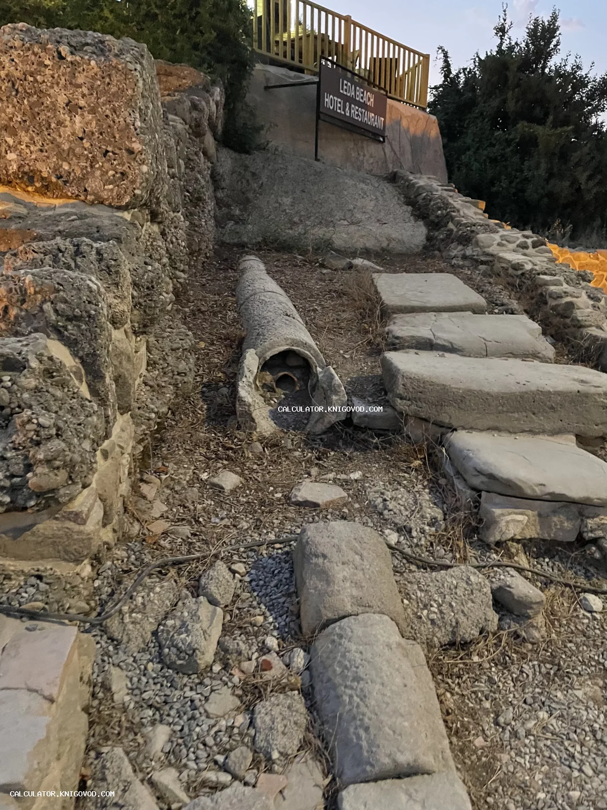 Древние каменные трубы и руины водопроводной системы на фоне современной вывески отеля.