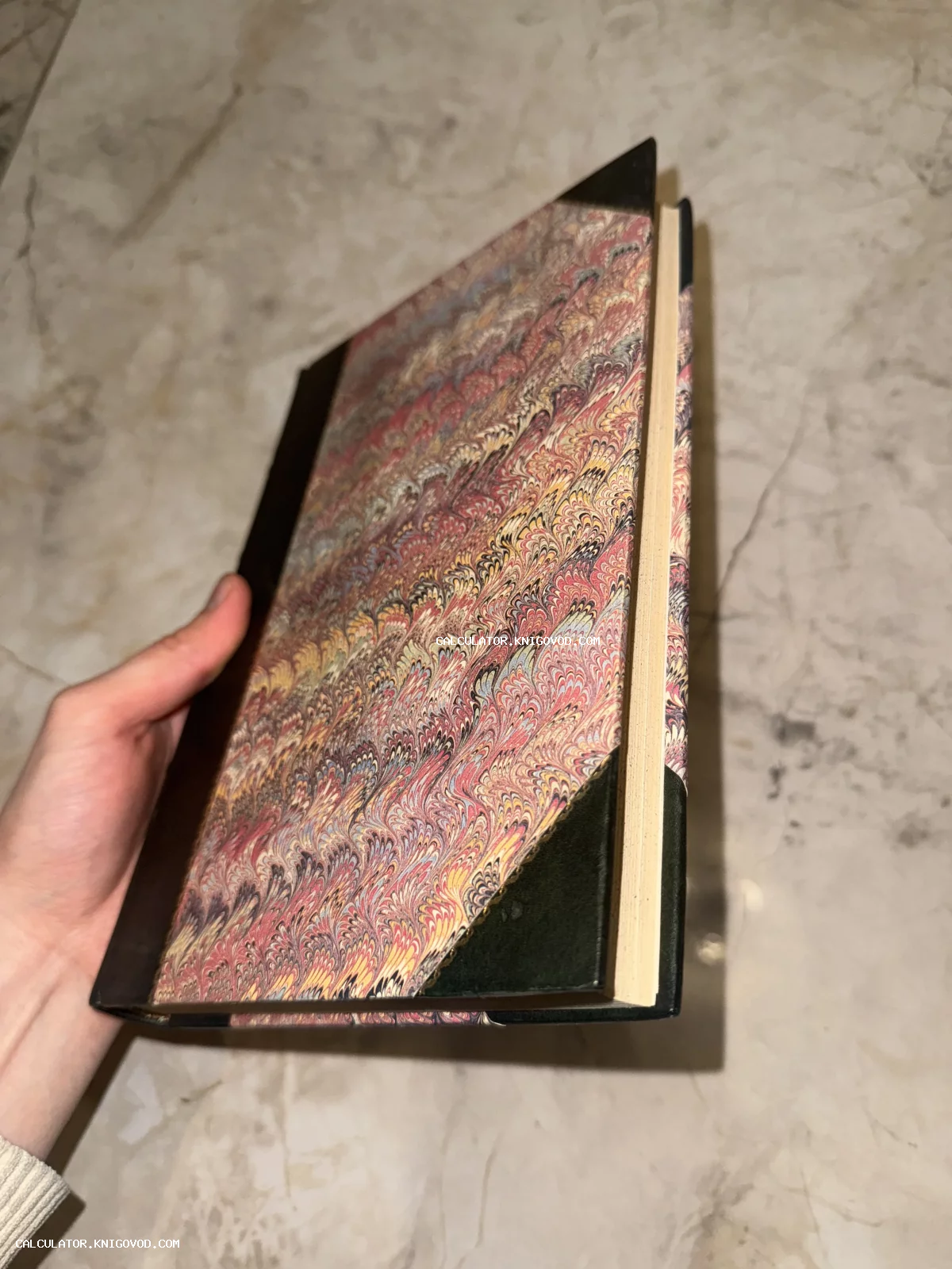 Старинная книга с обложкой из цветной мраморной бумаги и кожаным корешком в руке.