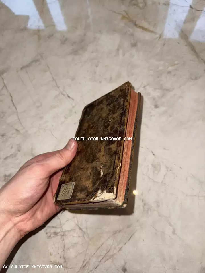 Мужская рука держит небольшую старинную книгу с сильно потертой обложкой и красным обрезом страниц на мраморном фоне.
