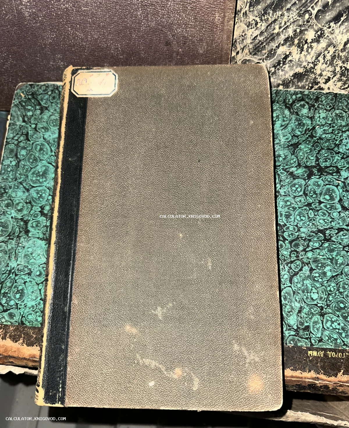 Старинная книга с серым фактурным переплетом, кожаным корешком и бумажным ярлыком на фоне других антикварных томов.