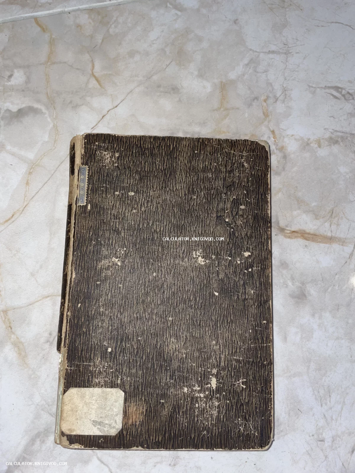 Вид сверху на обложку старинной книги с темным узором под дерево, потертостями по краям и бумажной наклейкой в нижнем углу.