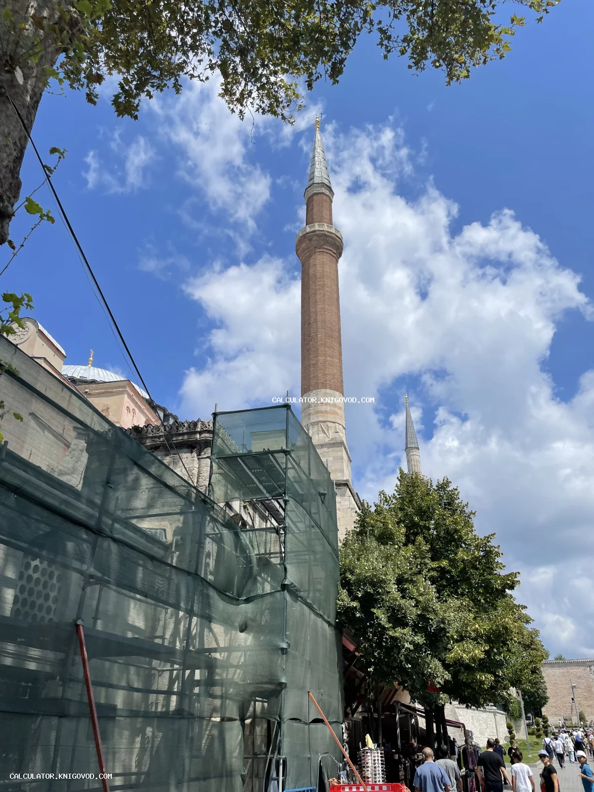 Высокий кирпичный минарет мечети Айя-София на фоне голубого неба с облаками, нижняя часть здания скрыта за строительными лесами с зеленой защитной сеткой.
