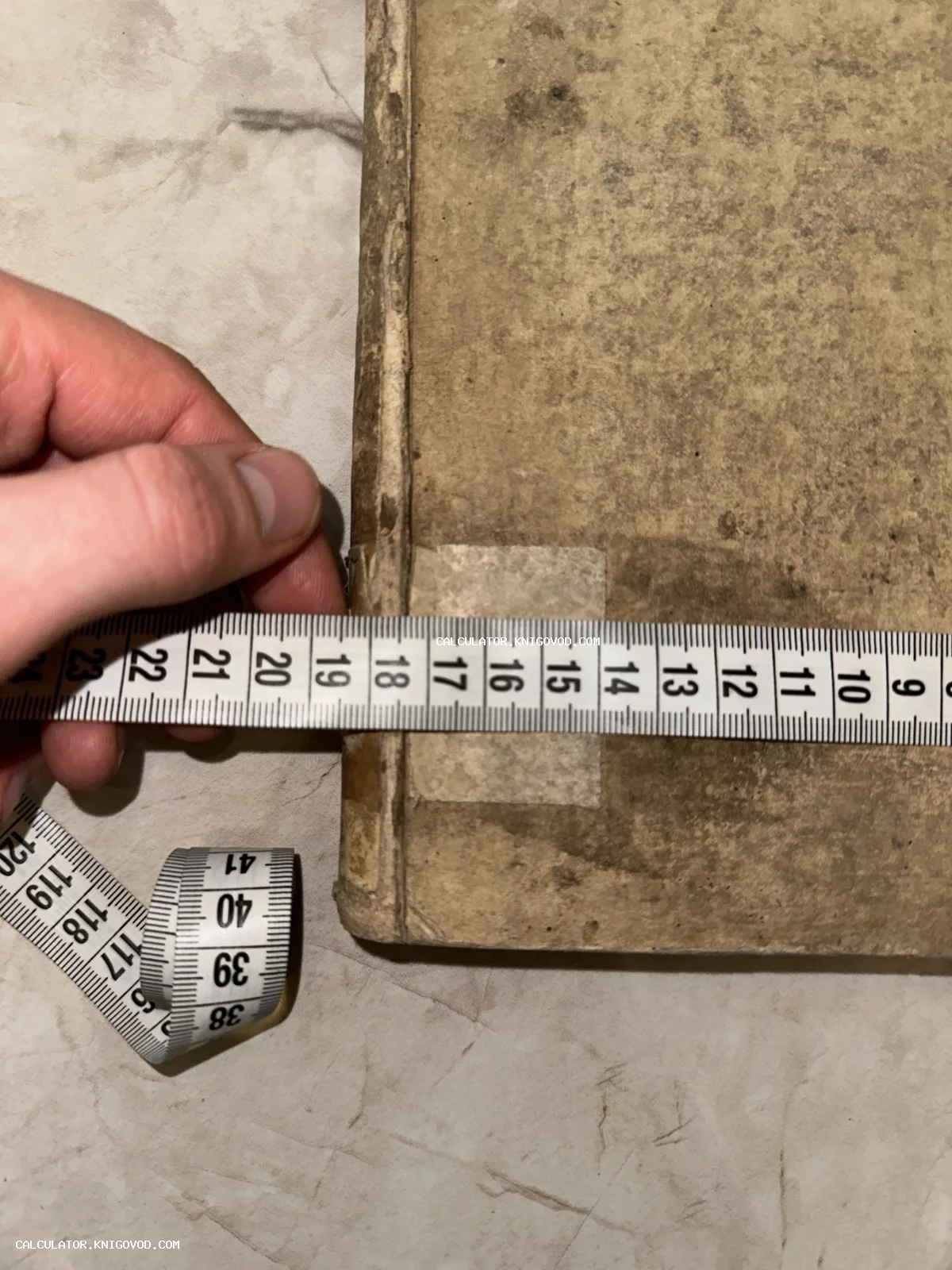 Рука измеряет ширину старинной книги в потрепанном бумажном переплете с помощью сантиметровой ленты.