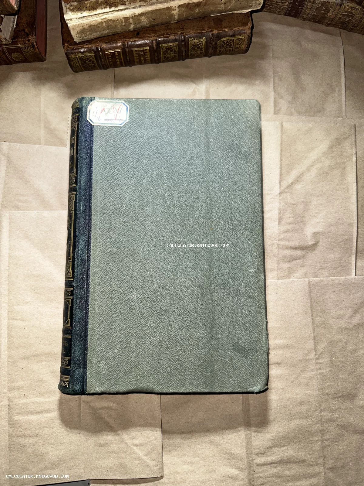 Старинная книга с бирюзовой обложкой и золотым тиснением на корешке на фоне крафтовой бумаги.