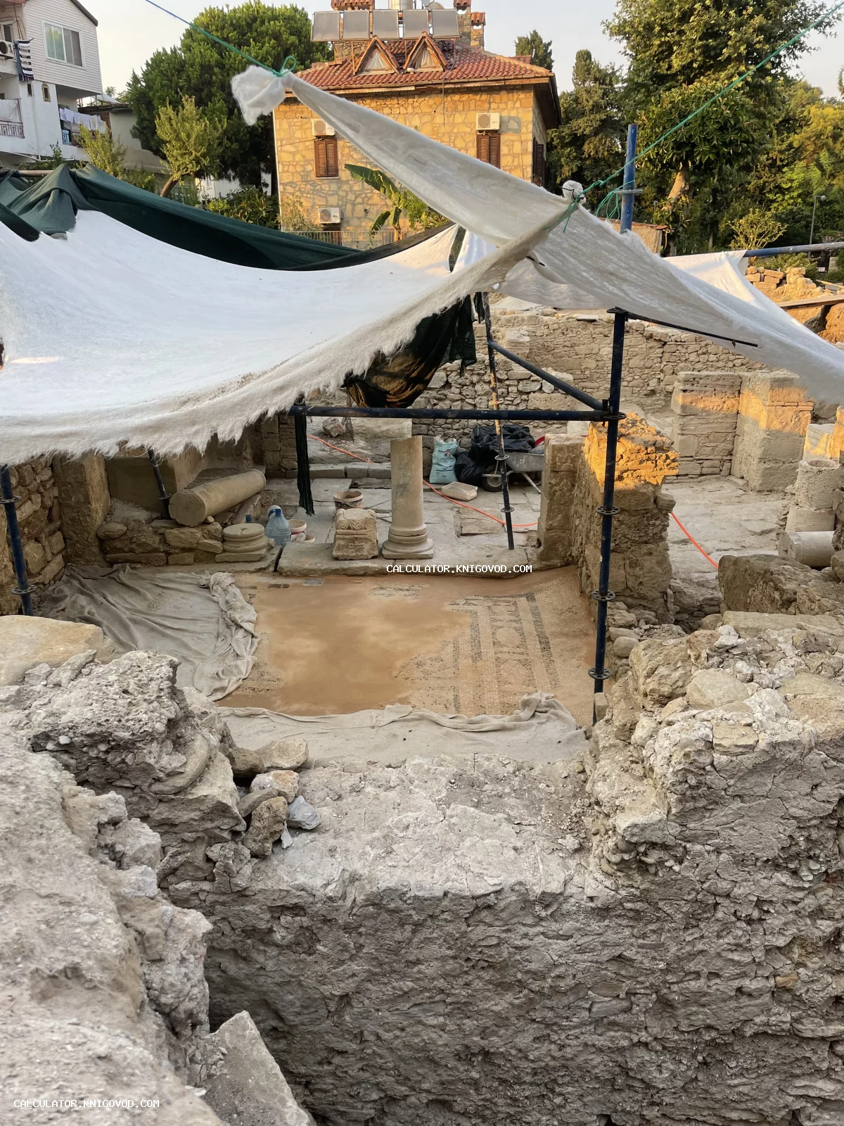 Место археологических раскопок с фрагментами античных колонн, каменными стенами и мозаичным полом под защитным белым навесом.