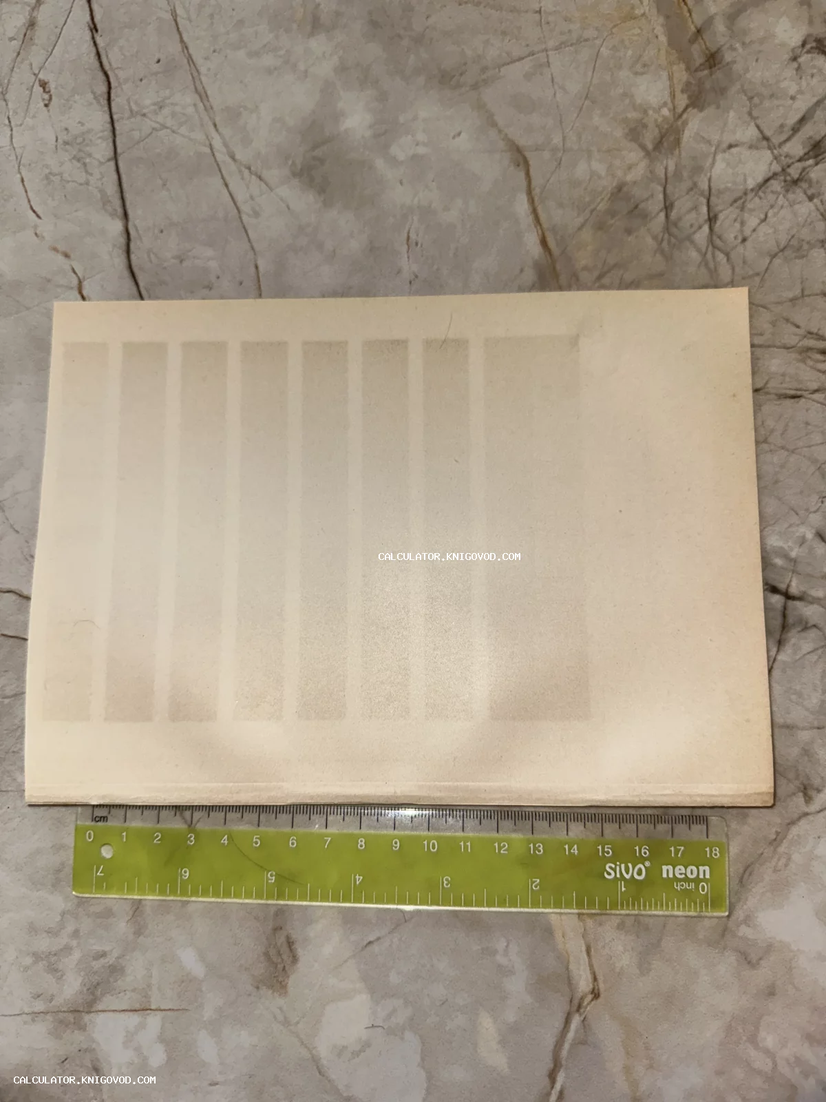Старинный пожелтевший лист бумаги с восемью вертикальными полосами, лежащий на мраморной поверхности рядом с зеленой пластиковой линейкой.