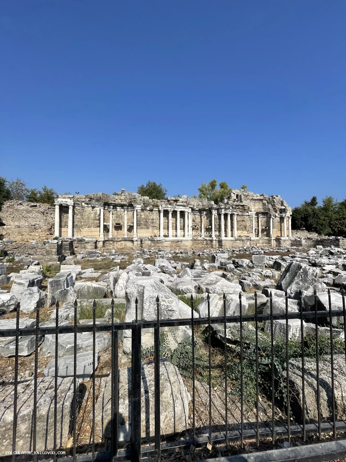 Руины античного монументального фонтана Нимфеум в Сиде с сохранившимися белыми колоннами и каменными блоками на фоне яркого синего неба.