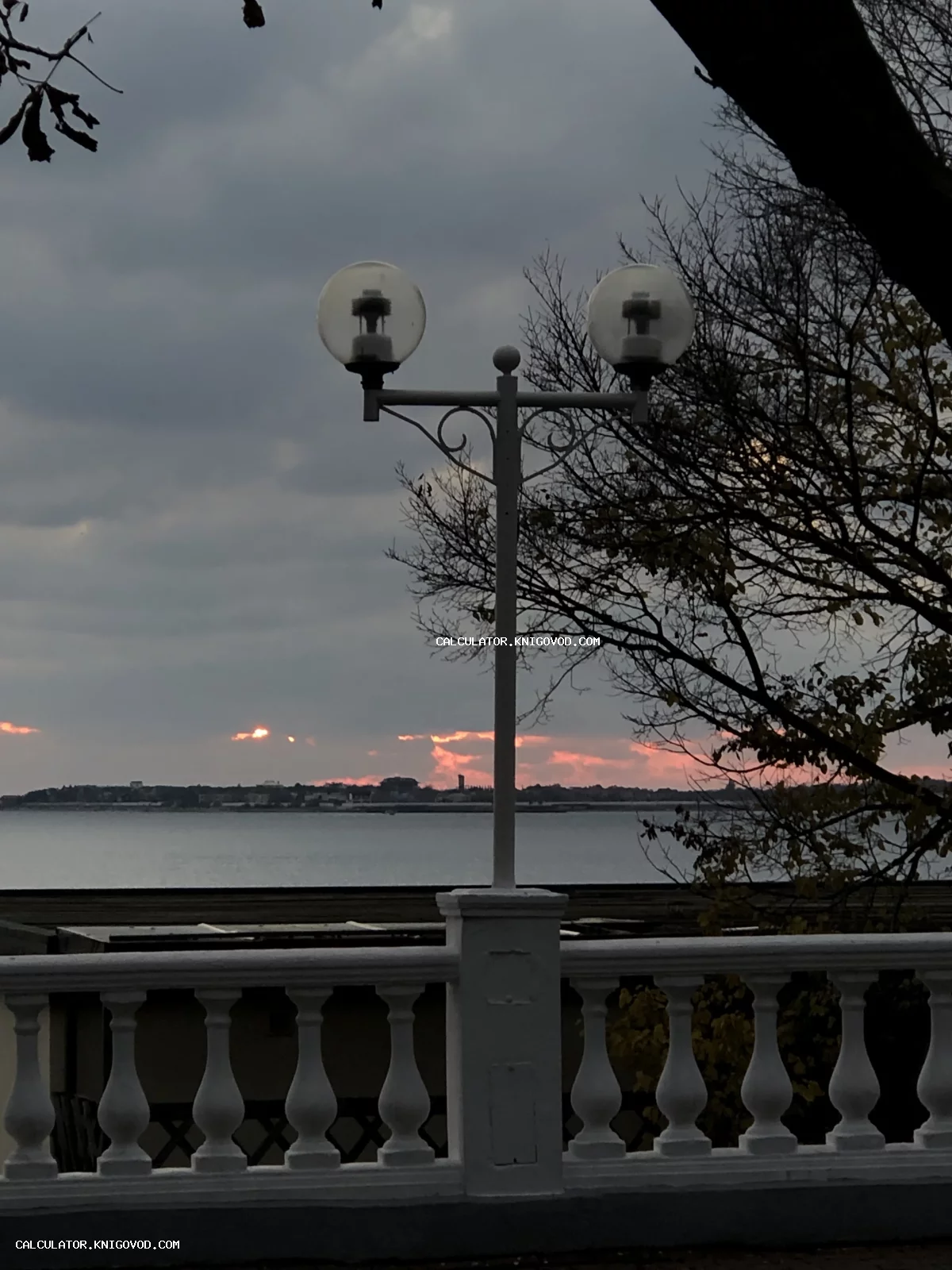 Классический уличный фонарь с двумя шарообразными плафонами на фоне вечернего моря и розового заката за белой балюстрадой.
