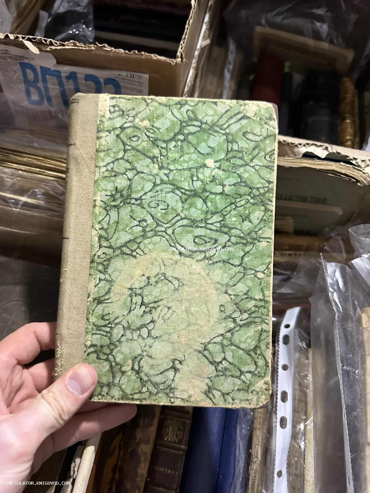 Рука держит старую книгу с зеленой мраморной обложкой и тканевым корешком на фоне коробок с архивными материалами.