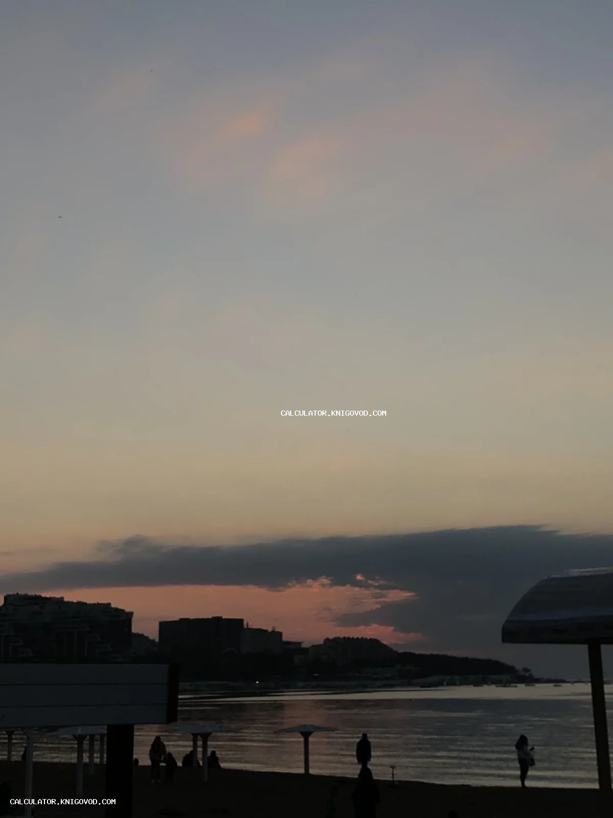 Пейзаж с темными силуэтами зданий и пляжных зонтов на фоне розового заката над морем.