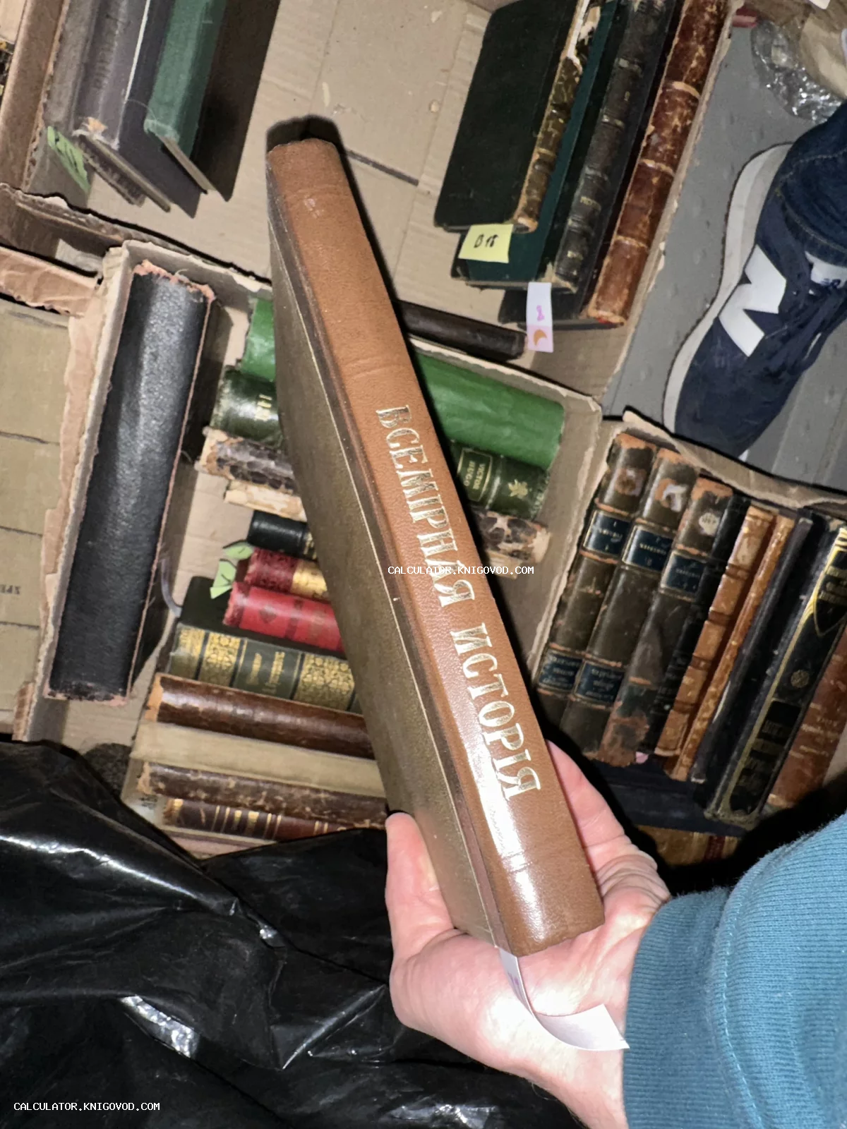 Старинная книга с золотым тиснением «Всемирная история» на корешке в руках человека на фоне коробок с другими книгами.
