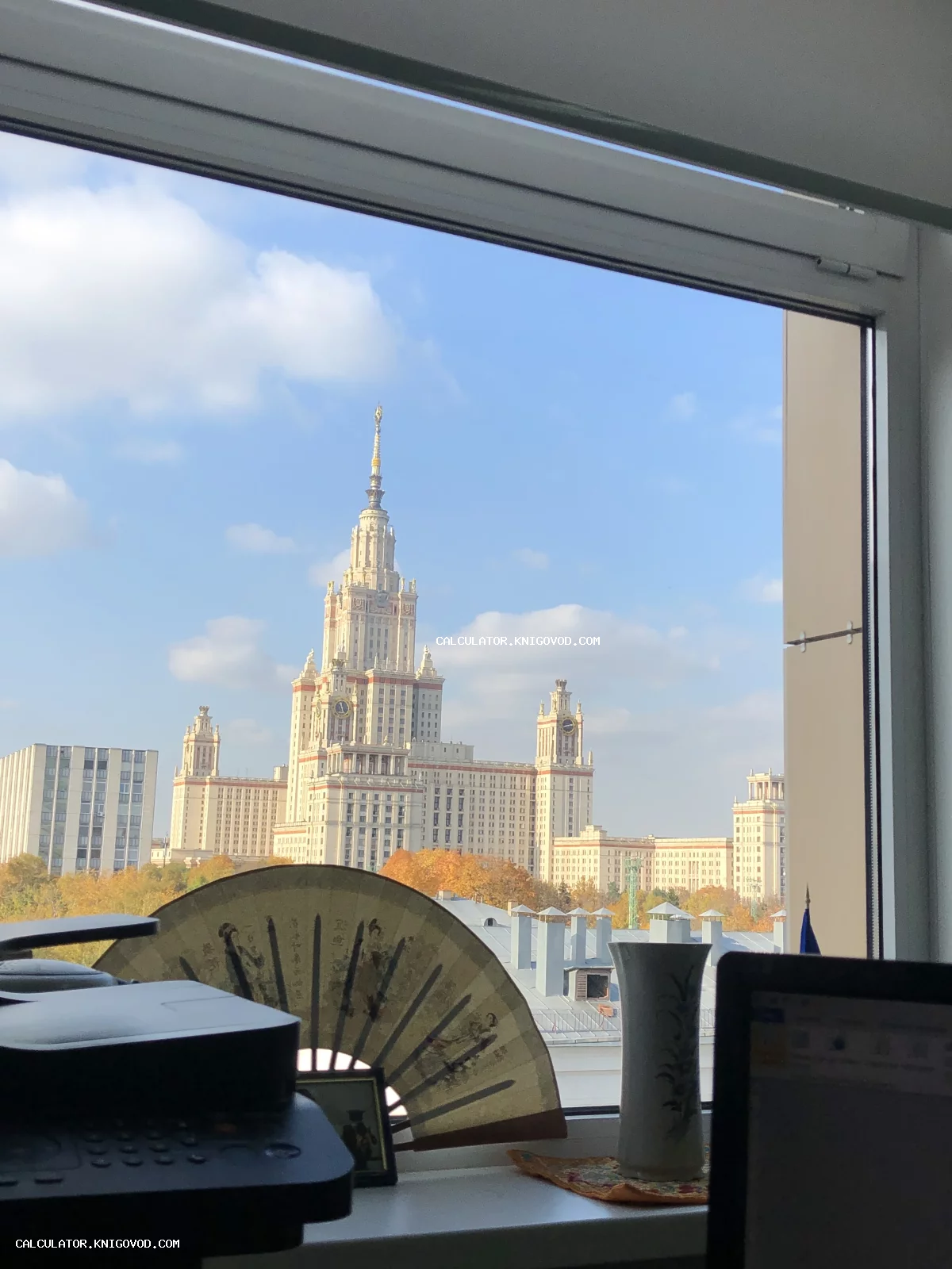 Вид из окна на сталинскую высотку МГУ в Москве осенью, на подоконнике лежит декоративный веер и стоит ваза.