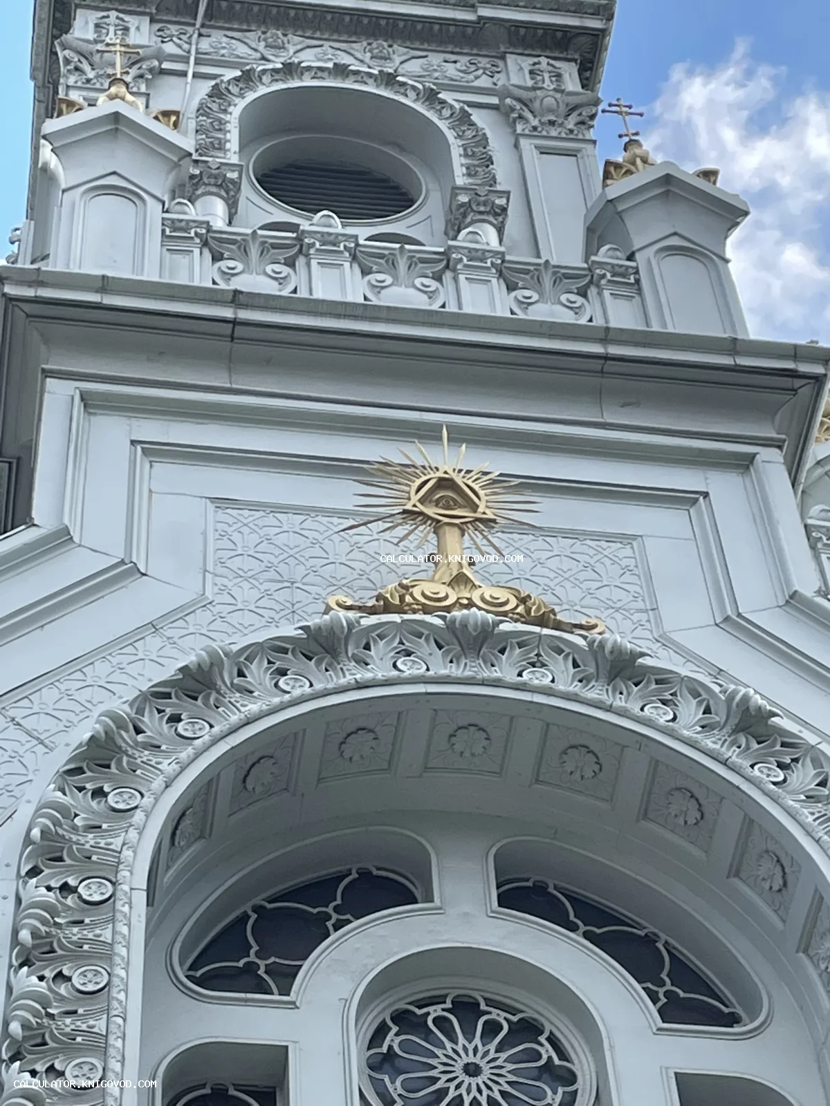 Фрагмент белого фасада здания с богатой лепниной и золотым символом Всевидящего ока в треугольнике над аркой.