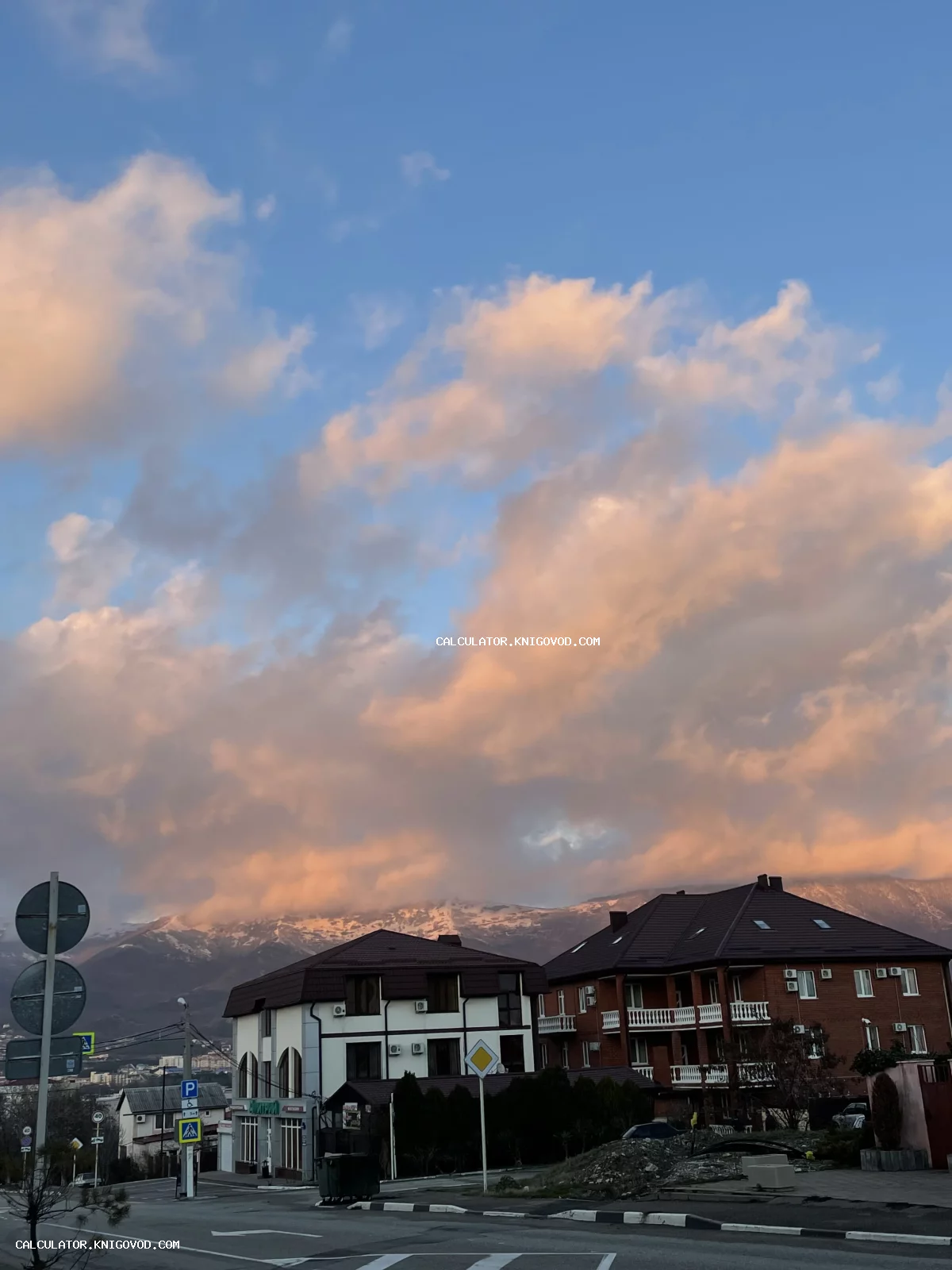 Фотография городских домов и улицы на фоне заснеженной горной гряды и облачного закатного неба.
