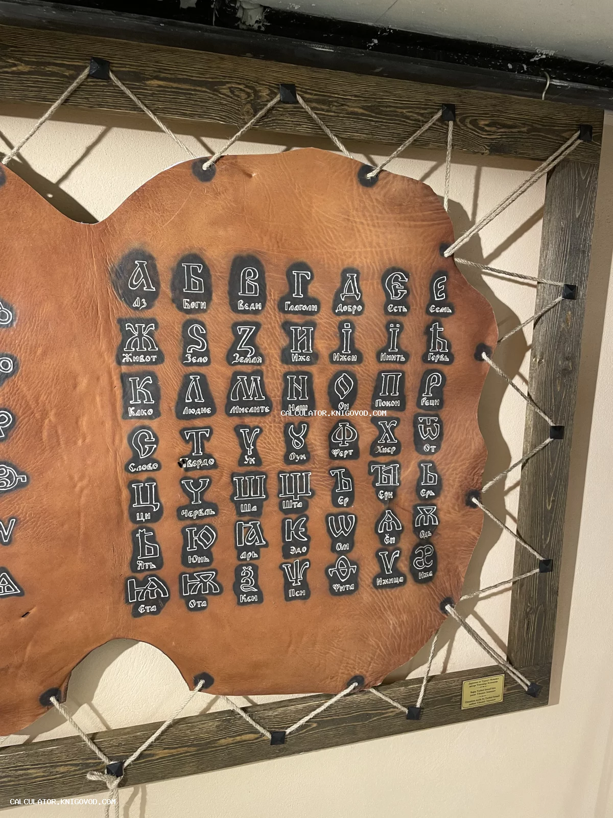 Декоративное настенное панно с буквами старославянского алфавита, нанесенными на кусок коричневой кожи, закрепленный веревками в деревянной раме.