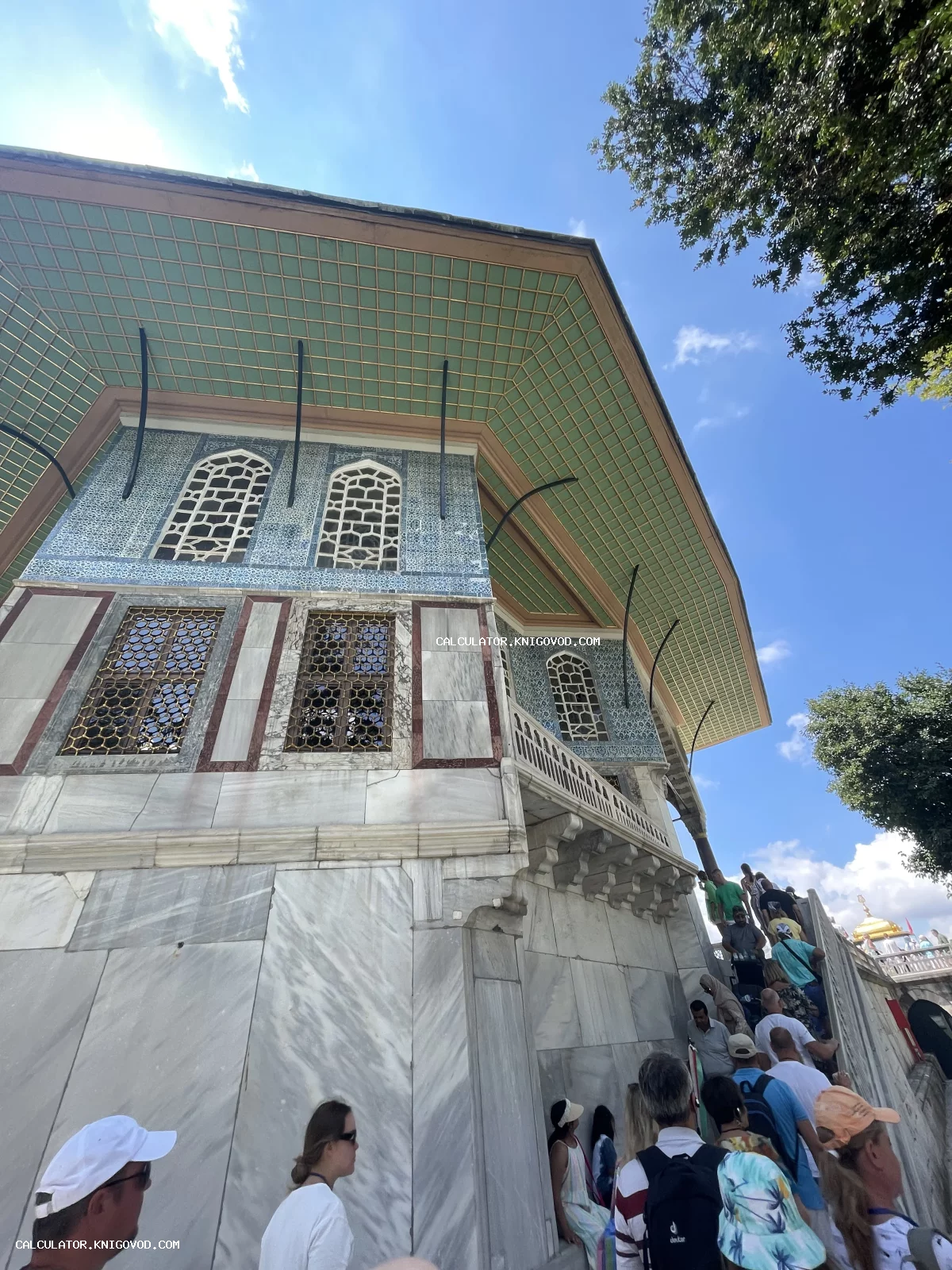 Вид снизу на фасад античного османского здания с голубой мозаикой, мраморными стенами и золочеными решетками на окнах под ярко-синим небом.