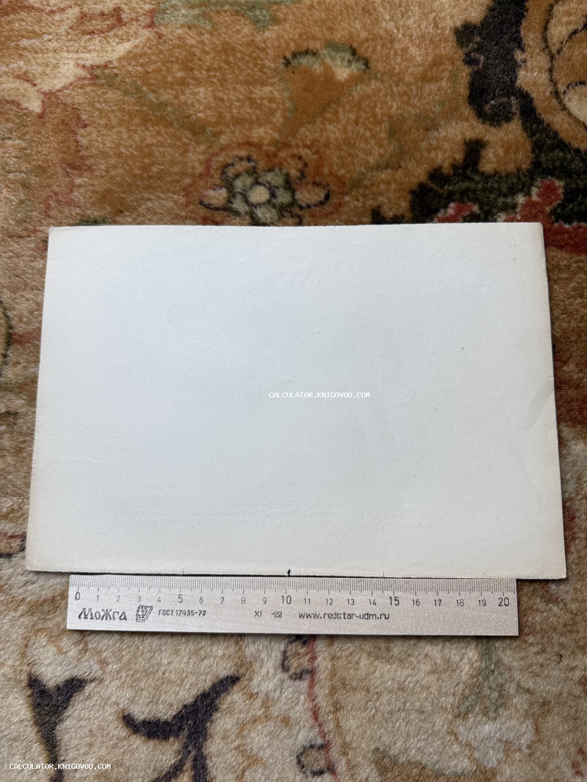 Чистая оборотная сторона старинной почтовой карточки на фоне ковра с приложенной деревянной линейкой для масштаба.
